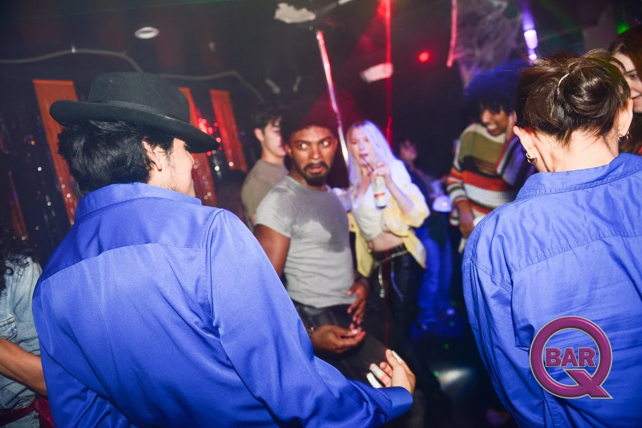 People dancing and socializing in a dimly lit nightclub with colorful lights and a 'Bar Q' logo in the corner.
