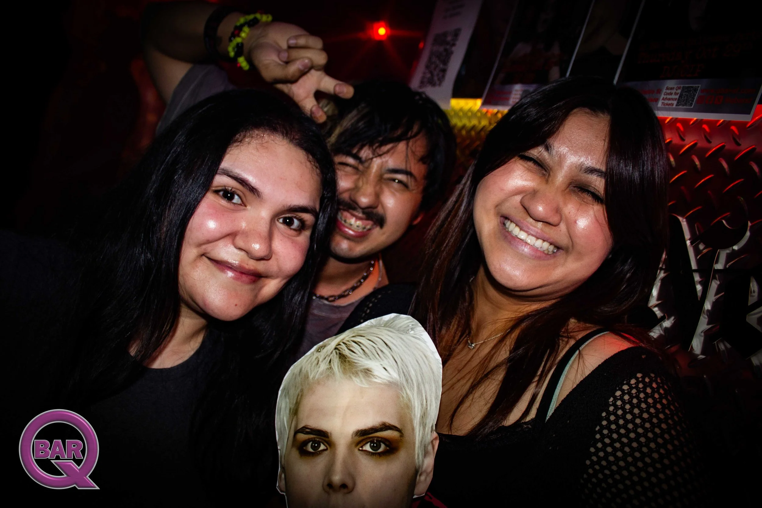 Group of smiling people in a dimly lit bar or club, with some holding tickets or wristbands, and a visible bar sign in the background.