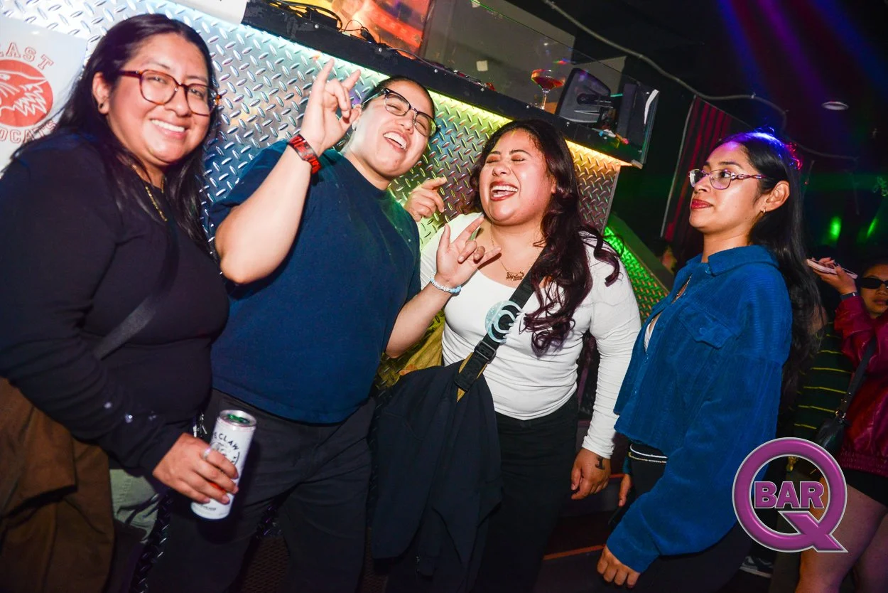 Four women smiling and enjoying themselves at a nightclub with colorful lights, one holding a can, two making hand gestures, and one wearing glasses.