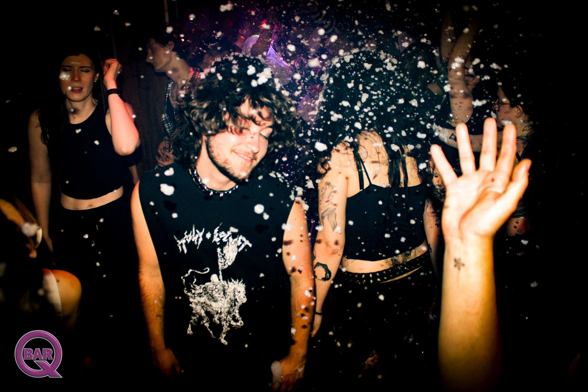 People dancing at a party with foam or snow falling around them, some with eyes closed and smiling, in a dark setting with party lights.