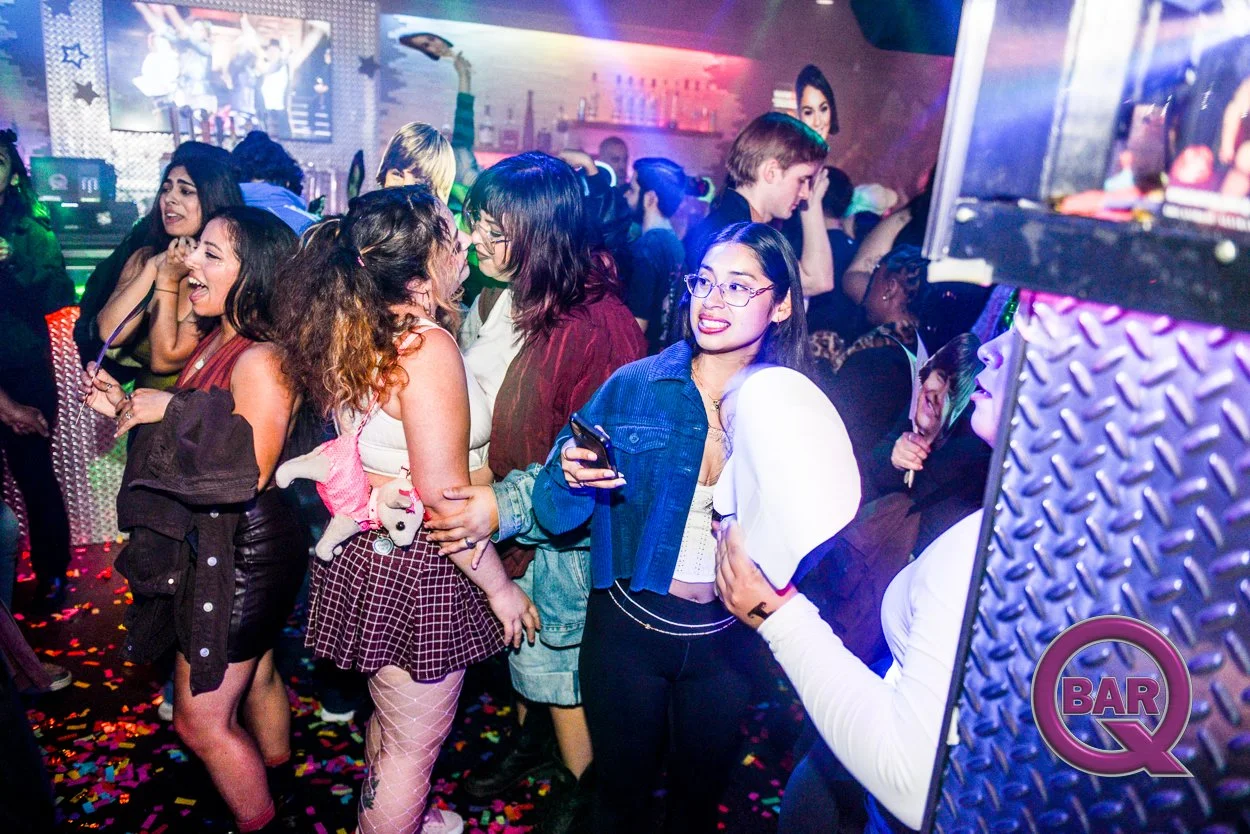 People dancing and socializing at a nightclub with colorful lights and confetti on the floor, with a DJ booth visible in the background.