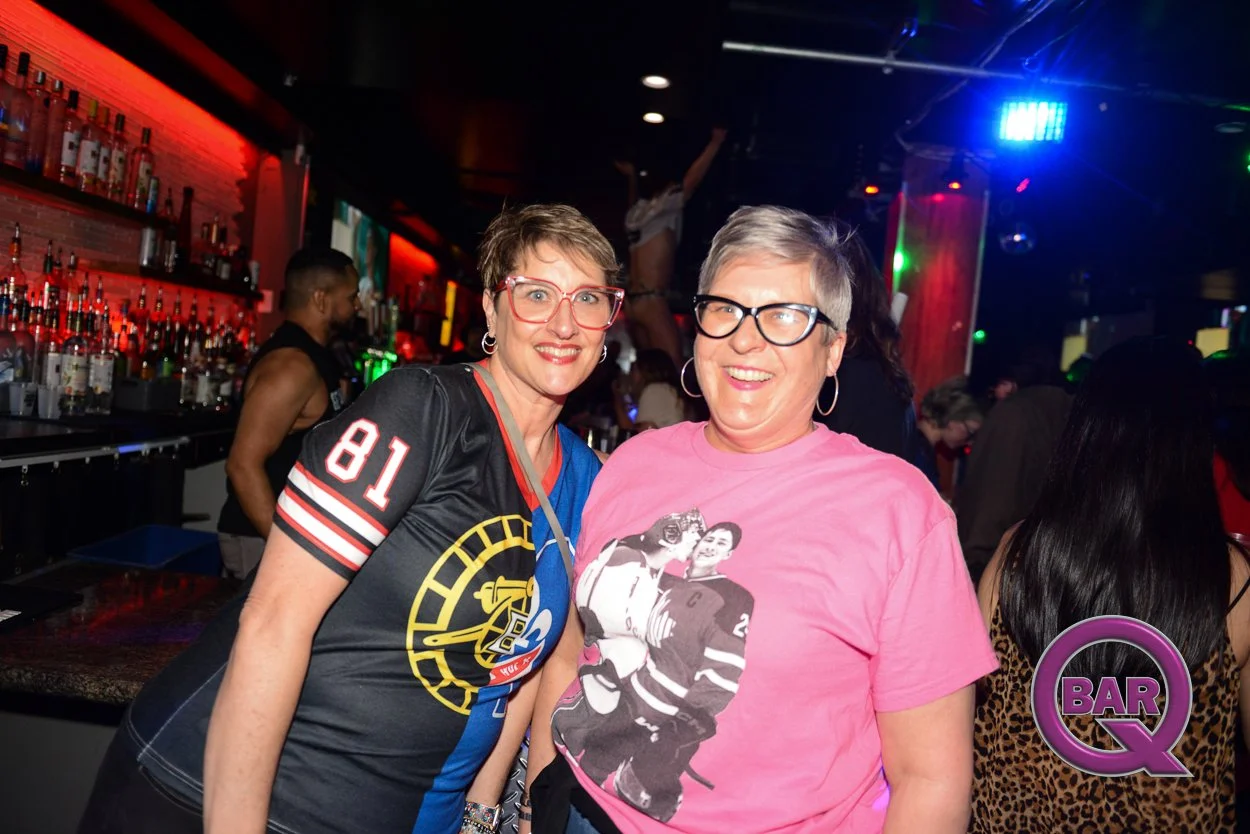 Two women smiling at a bar with colorful lights, one wearing a black sports jersey and the other a pink T-shirt with a hockey image, in a lively nightlife setting.