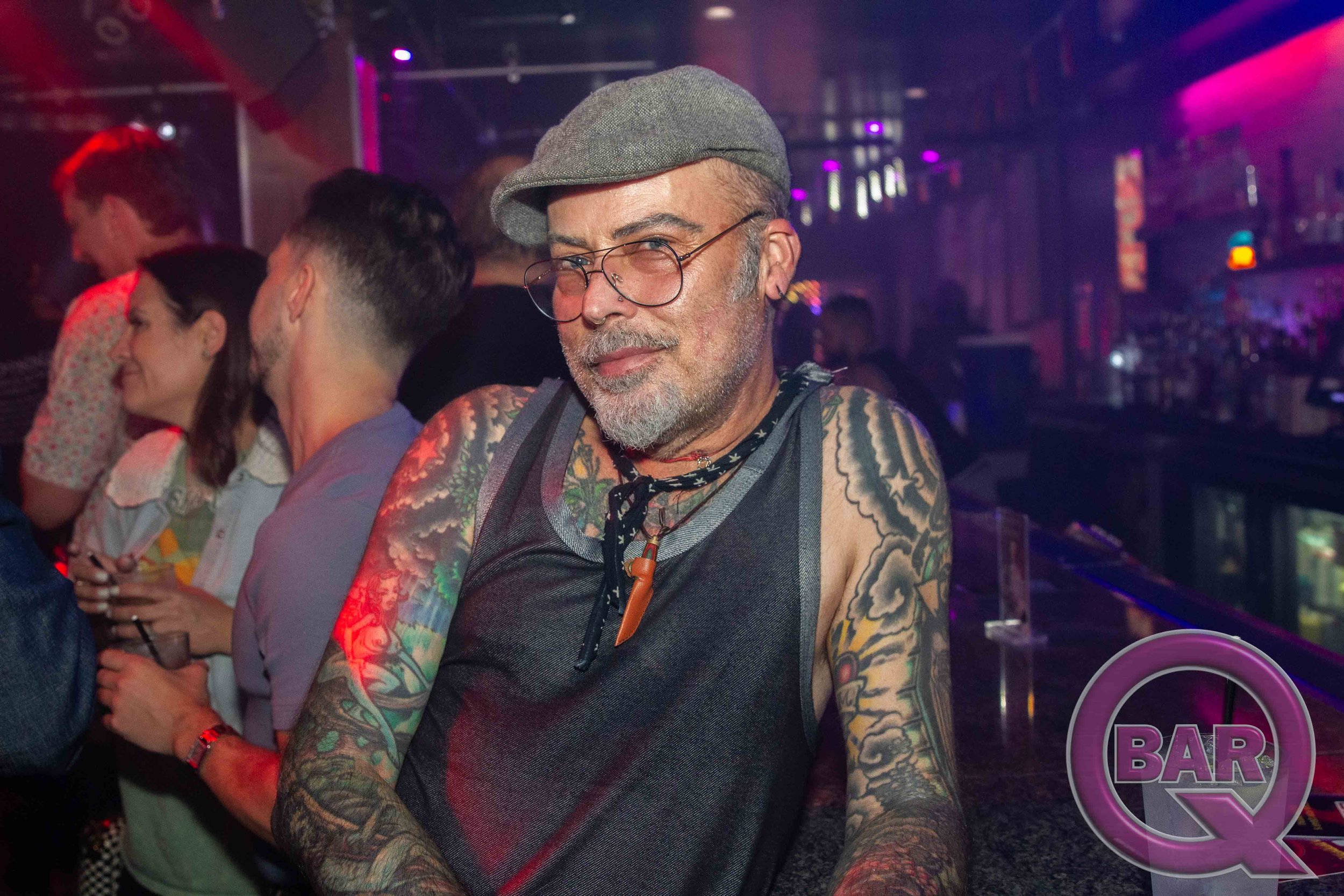 A man with tattoos, glasses, and a gray cap smiling at a bar at night, with other people in the background.