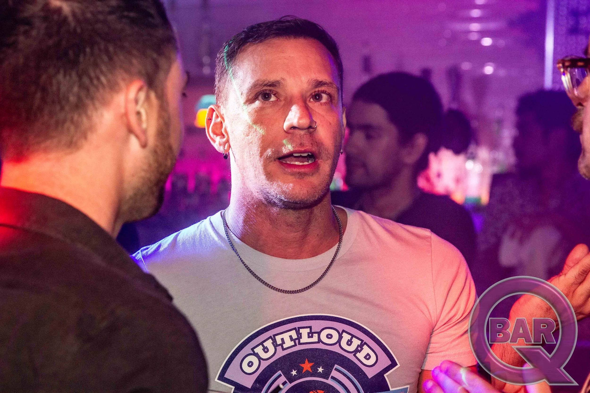 A man with short dark hair, earrings, and a nose ring is speaking to another person in a bar with purple lighting. The man is wearing a white t-shirt with a logo and a necklace.