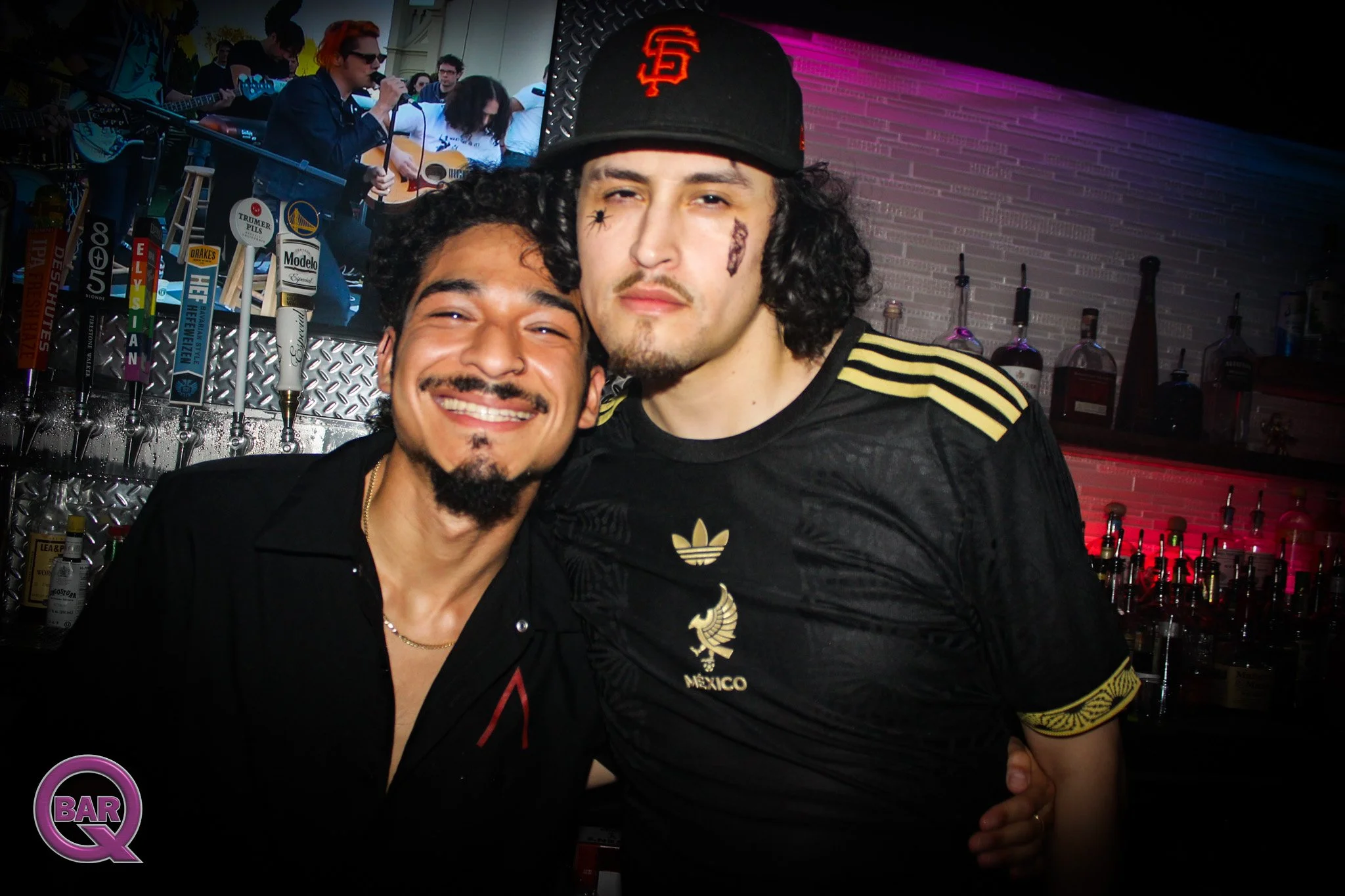 Two men smiling and posing for a photo at a bar. One man has dark curly hair and a goatee, wearing a black shirt. The other man has light curly hair, a mustache, and goatee, wearing a black Adidas jersey with gold accents and a cap with a red logo. I