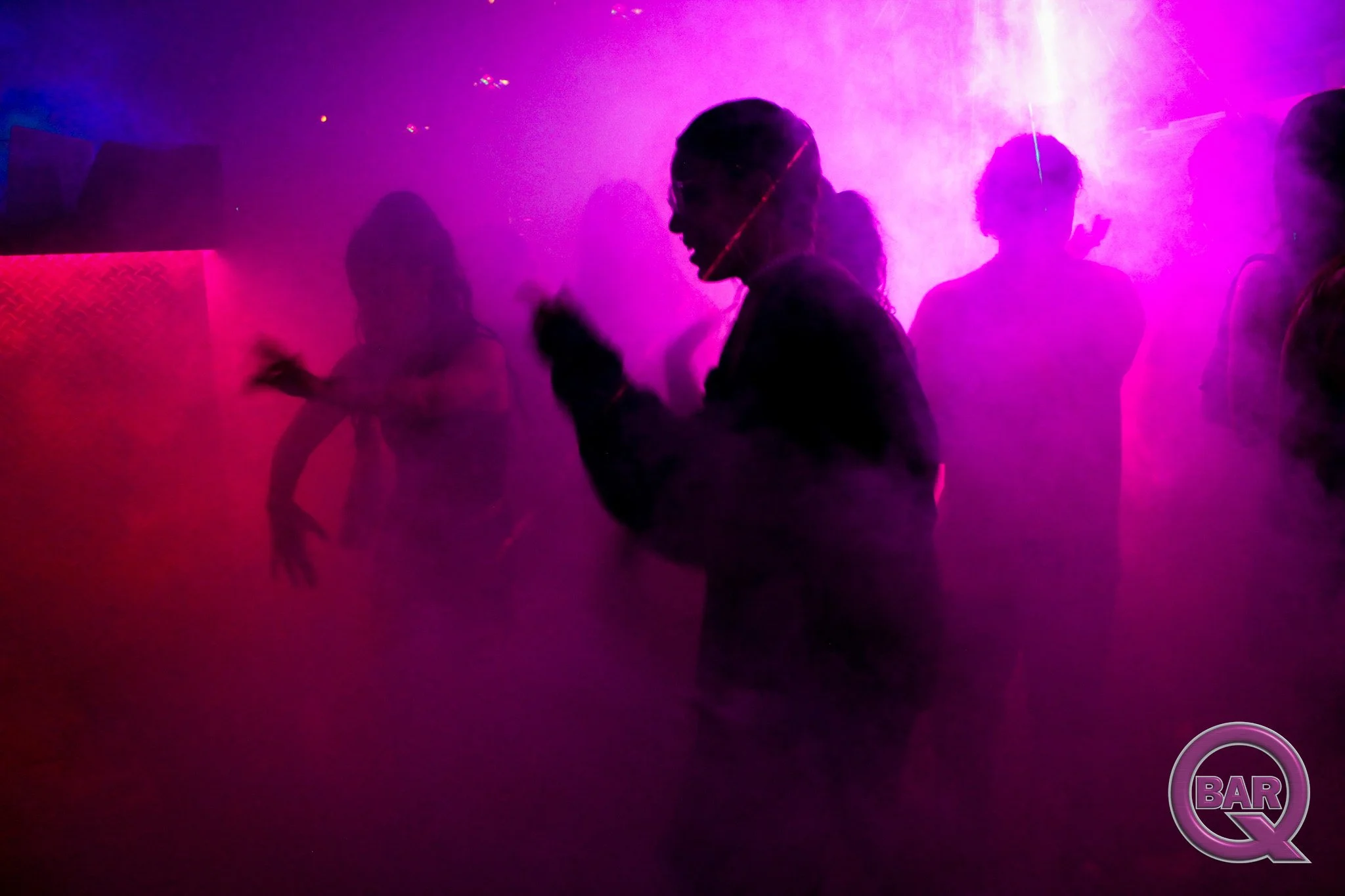 People dancing in a nightclub with purple and pink lighting and fog.