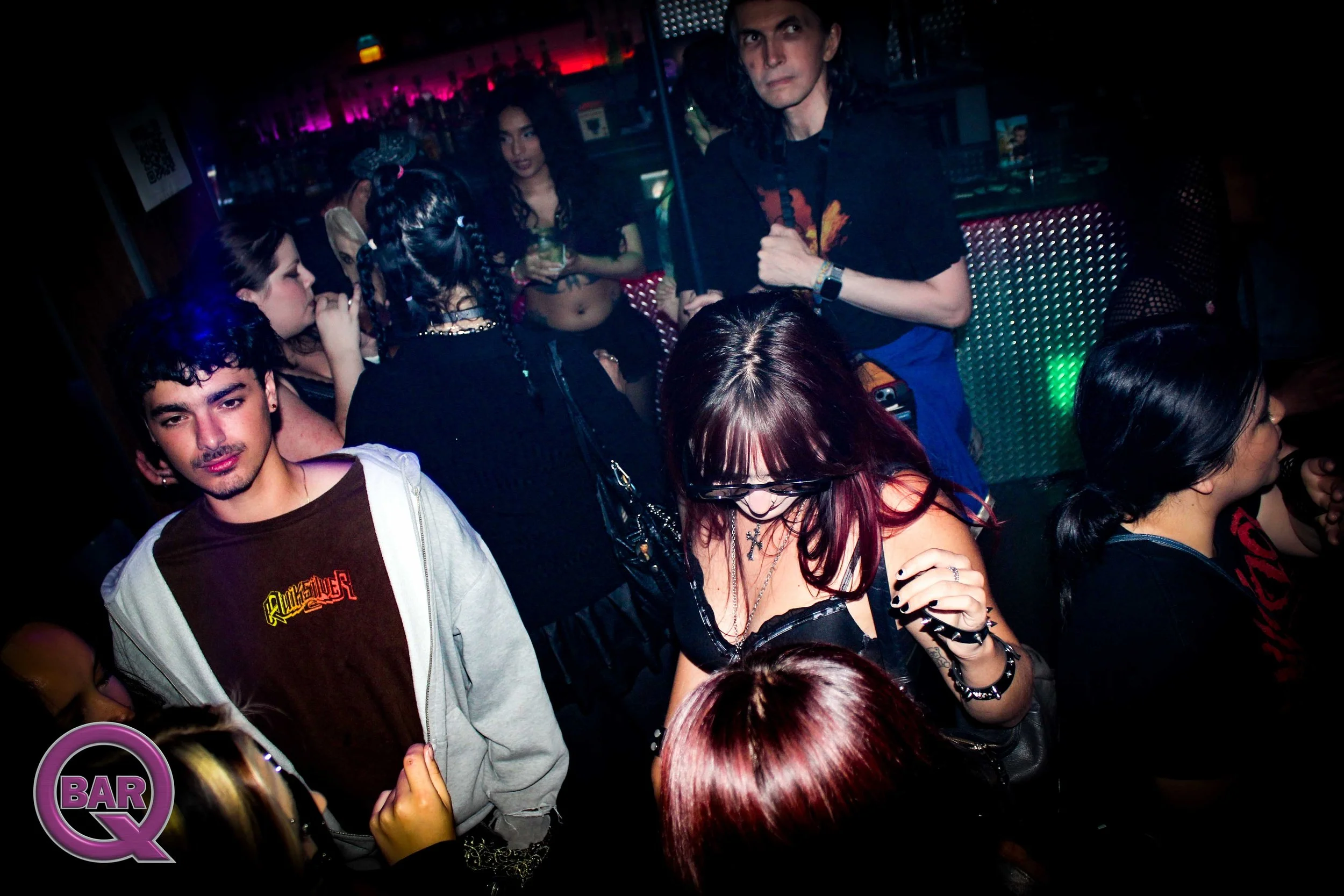 People socializing at a bar or nightclub with colorful lighting. Several individuals are engaged in conversation or relaxing, with one man standing holding something in his hand, and a woman with red hair and jewelry adjusting her bracelet in the for