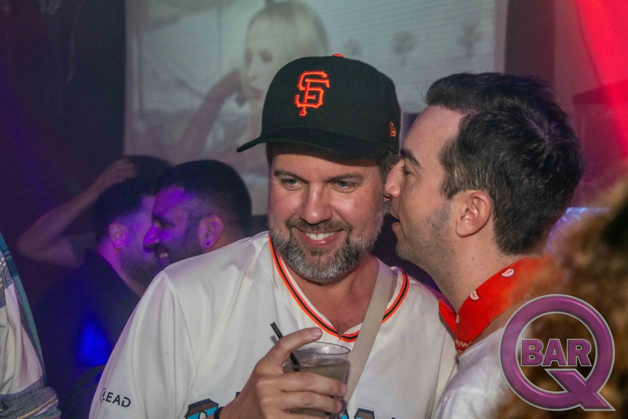 Two men are smiling and talking at a bar or party. One is wearing a black San Francisco Giants hat, and the other has dark hair and is wearing a red scarf with white patterns. The man with the hat is holding a drink.