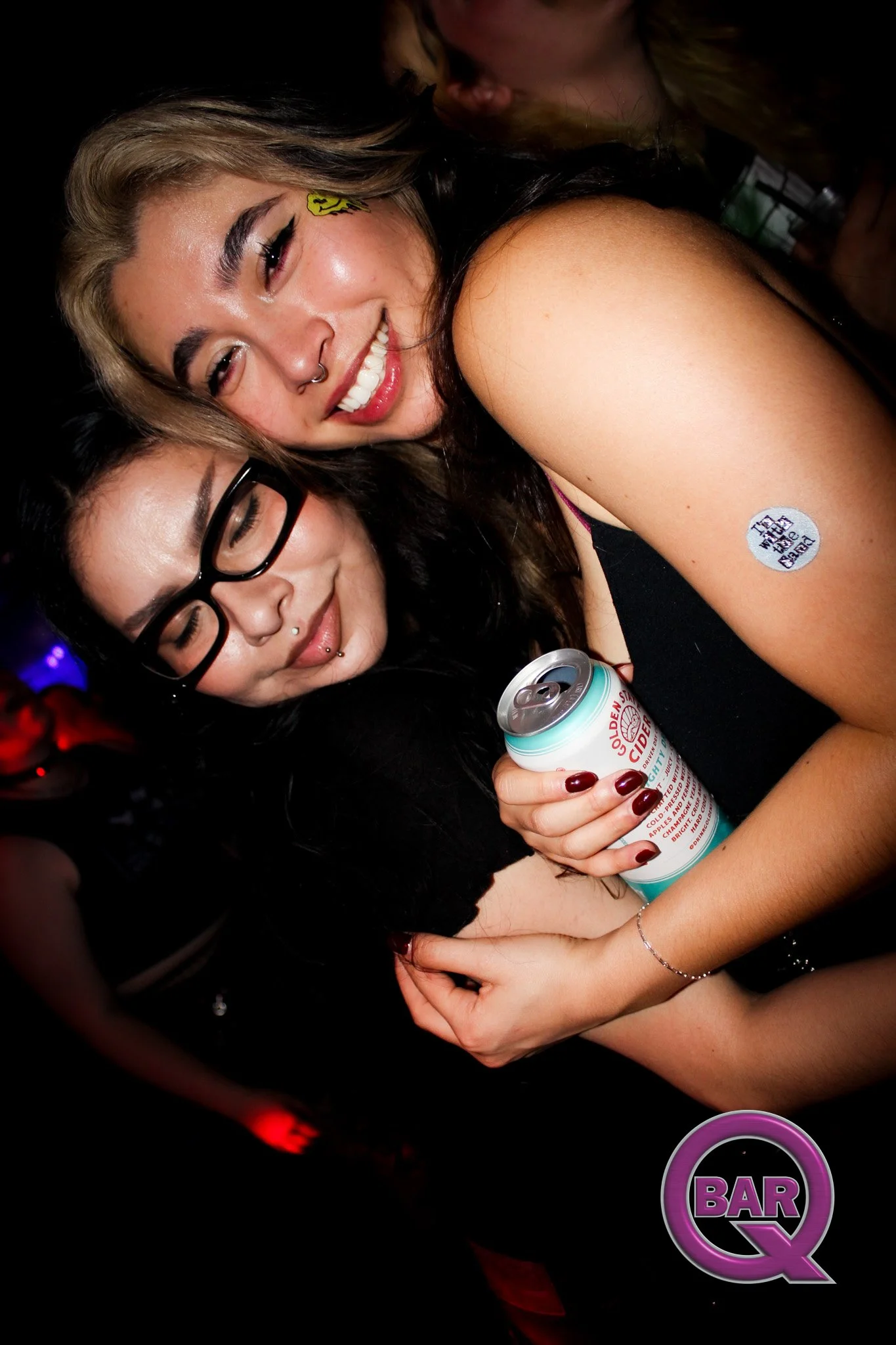 Two women smiling and hugging at a bar, with one holding a can of cider, in a dark setting with a neon bar sign visible.