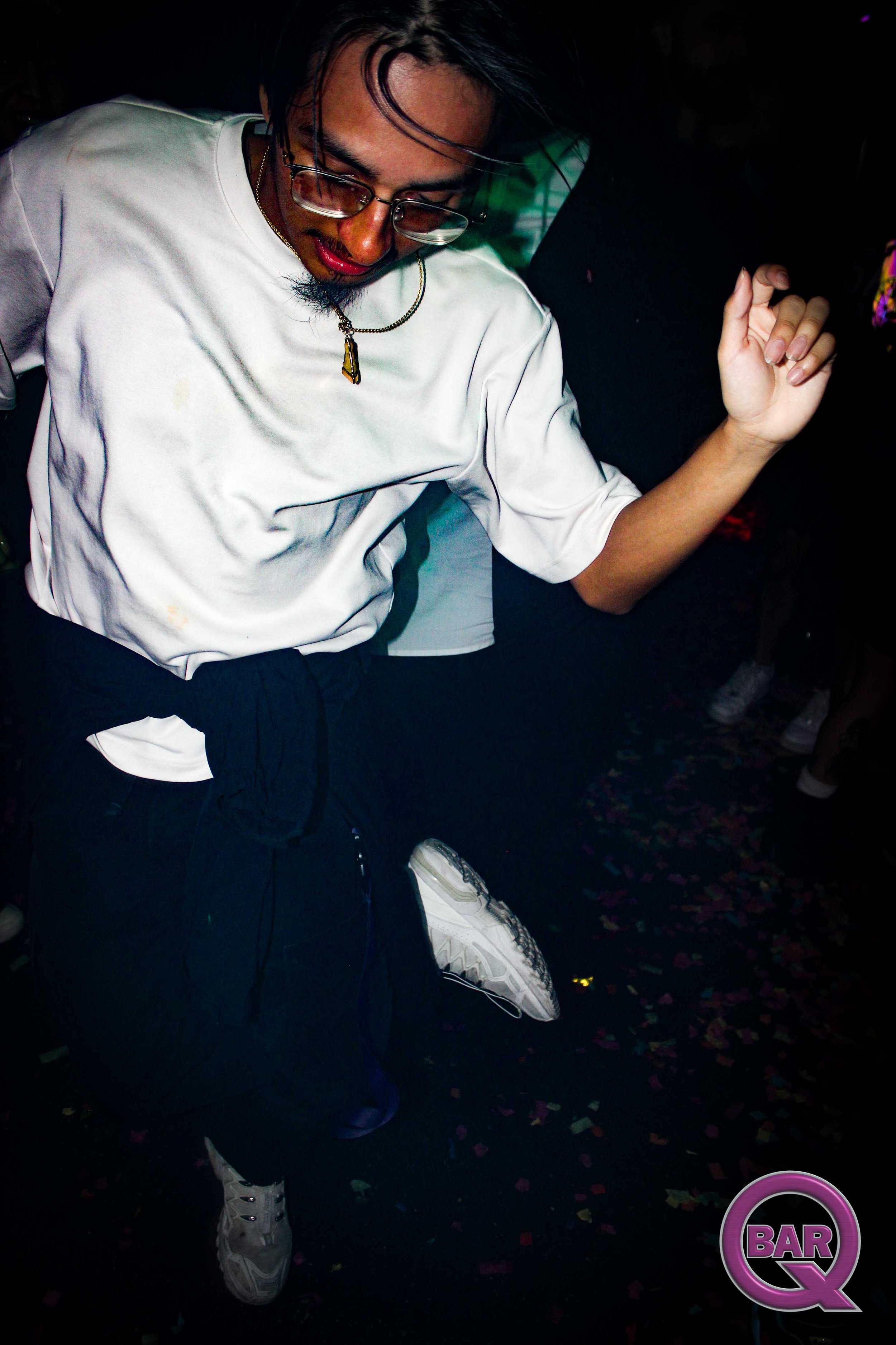 A person with long dark hair, glasses, and red lipstick dancing in a dark club with confetti on the floor and a purple "BAR Q" logo in the corner.