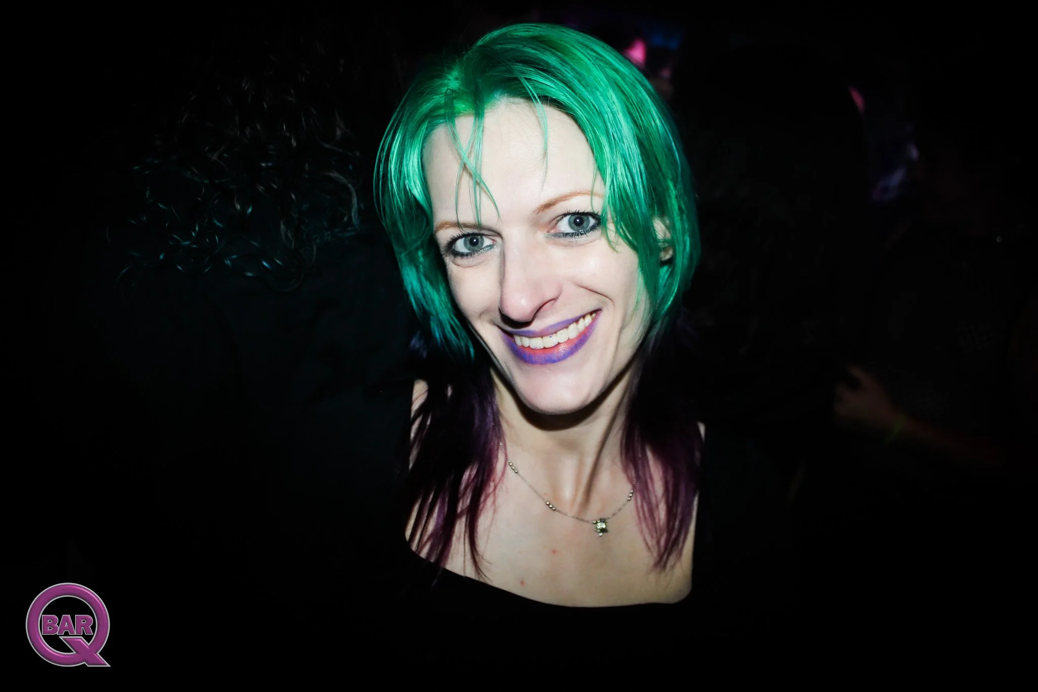 A woman with bright green hair, blue eyes, and purple lipstick smiling at the camera, wearing a black top and a necklace, in a dark environment with a small pink and purple 'Q BAR' logo in the bottom left corner.
