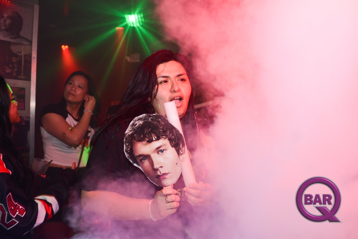 A woman holding a large cutout photograph of a man with curly hair at a party with colorful smoke and lights