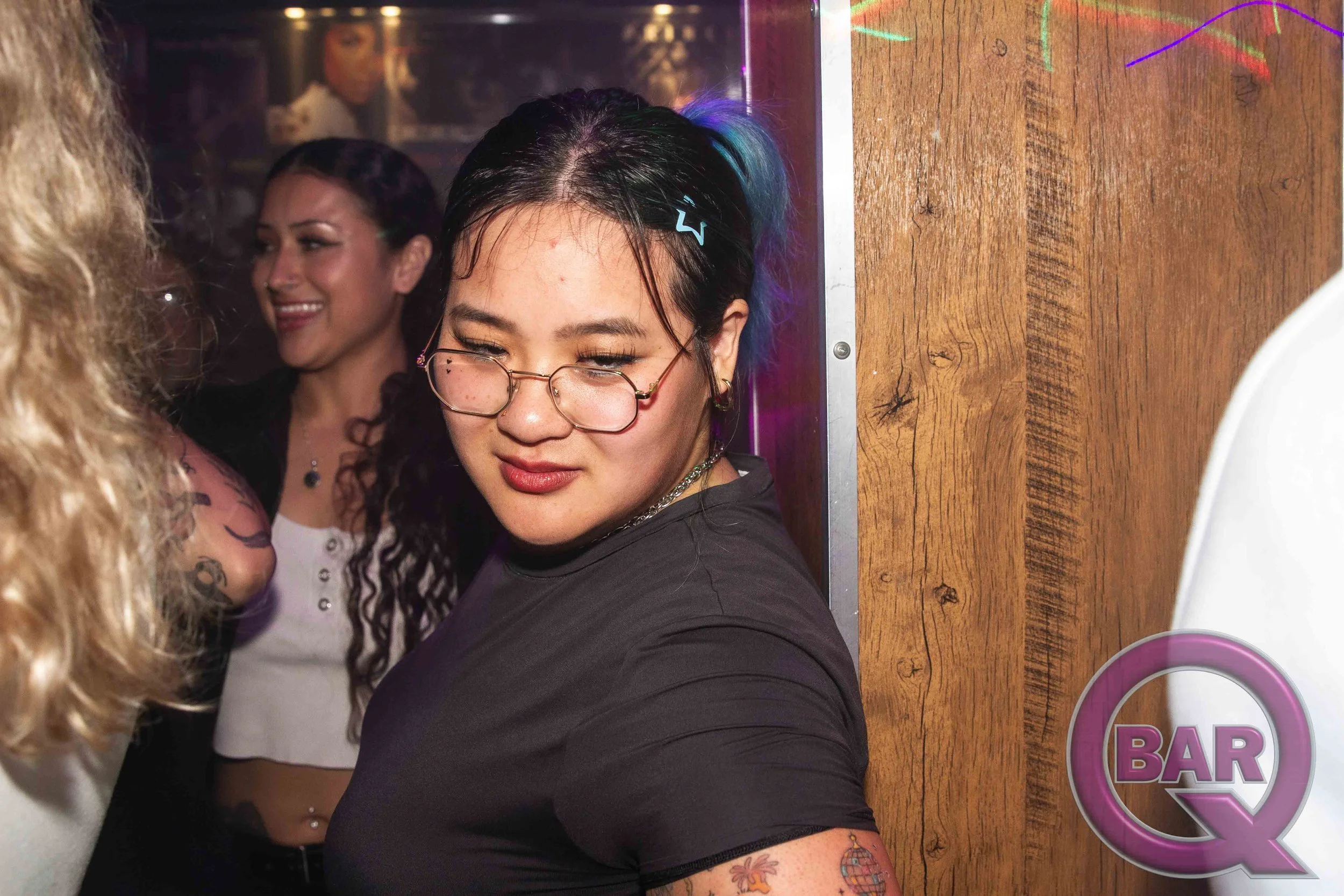 A group of people at a bar, including a woman with glasses and colorful hair, smiling and interacting with others.