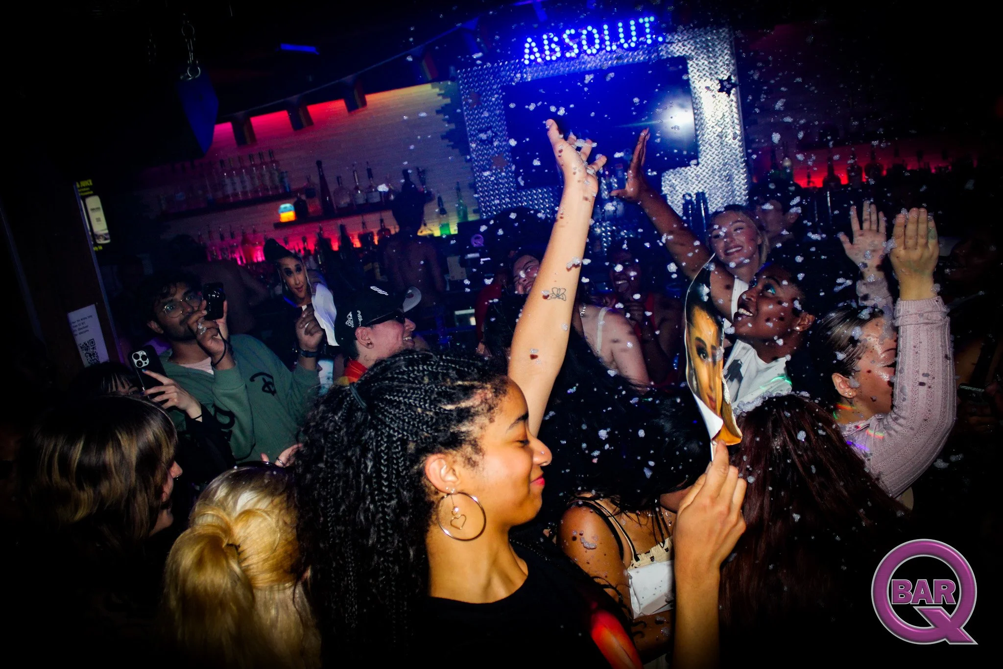 Group of people at a lively nightclub or bar, with a neon sign that reads 'ABSOLUTE,' surrounded by colorful lights and decorations, dancing and celebrating with one person's hands raised, confetti falling around.