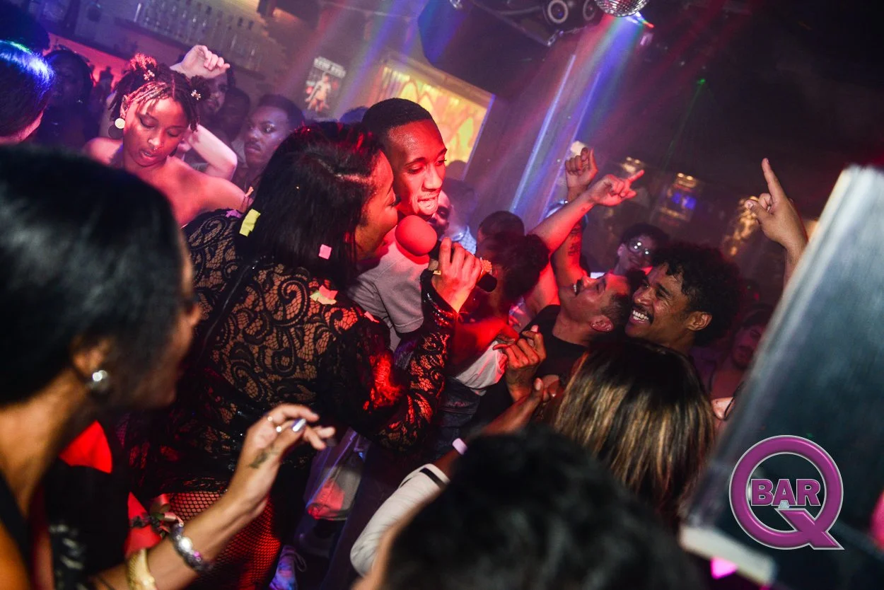 People dancing and singing at a lively nightclub with colorful lighting and a DJ. A woman is holding a microphone, and others are enjoying the music and celebration.