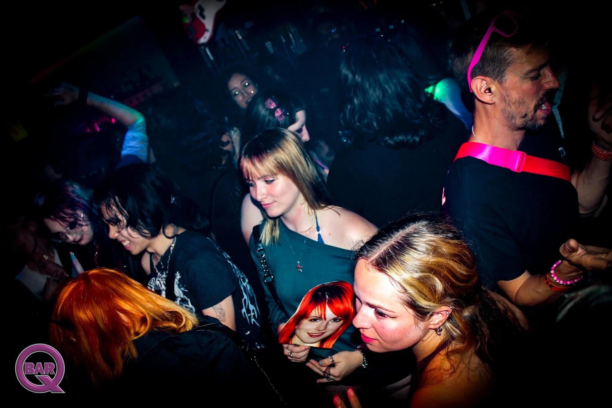 A crowded nightclub with people dancing and socializing, some are holding drinks and a poster of a woman with bright red hair is visible in the foreground.