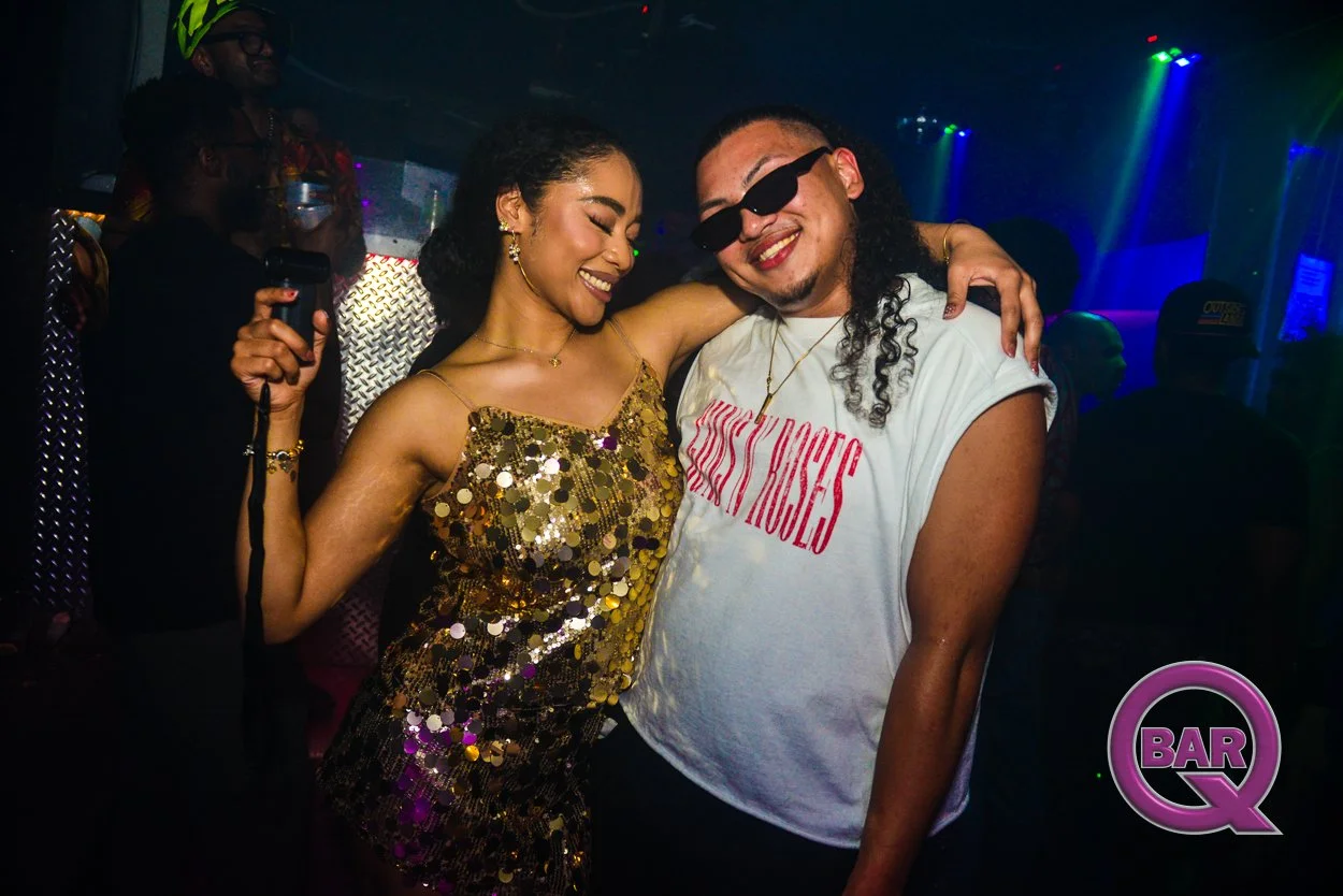 Two people smiling and hugging at a nightclub with colorful lighting. The woman wears a gold sequin dress, and the man wears sunglasses and a white T-shirt. There are other people in the background, and a purple 