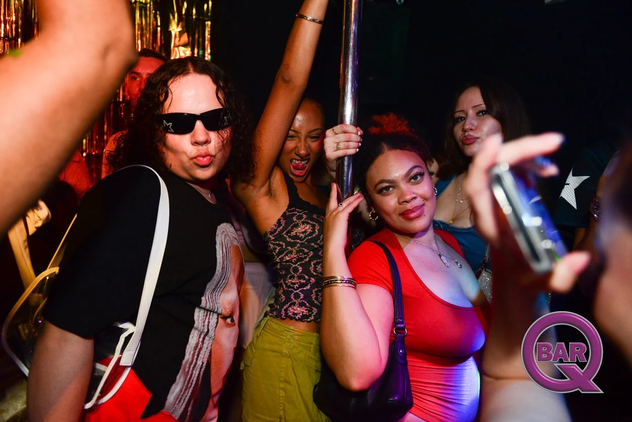 Group of women partying with one wearing sunglasses and making a kiss face, another holding a pole, and others posing for the camera at a nightclub.