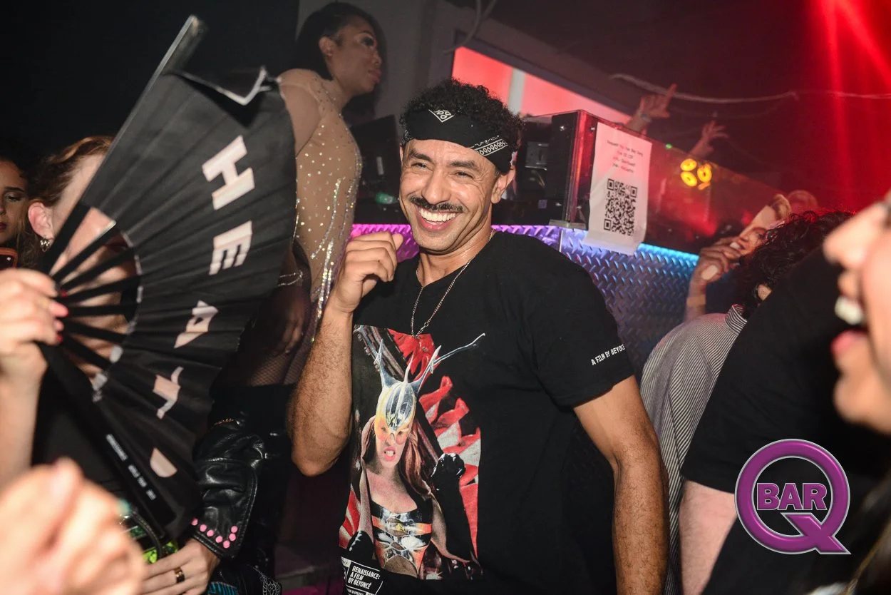 Young man with curly hair wearing a black bandana and a graphic t-shirt, smiling and enjoying a night out at a bar or club, surrounded by people and colorful lights.