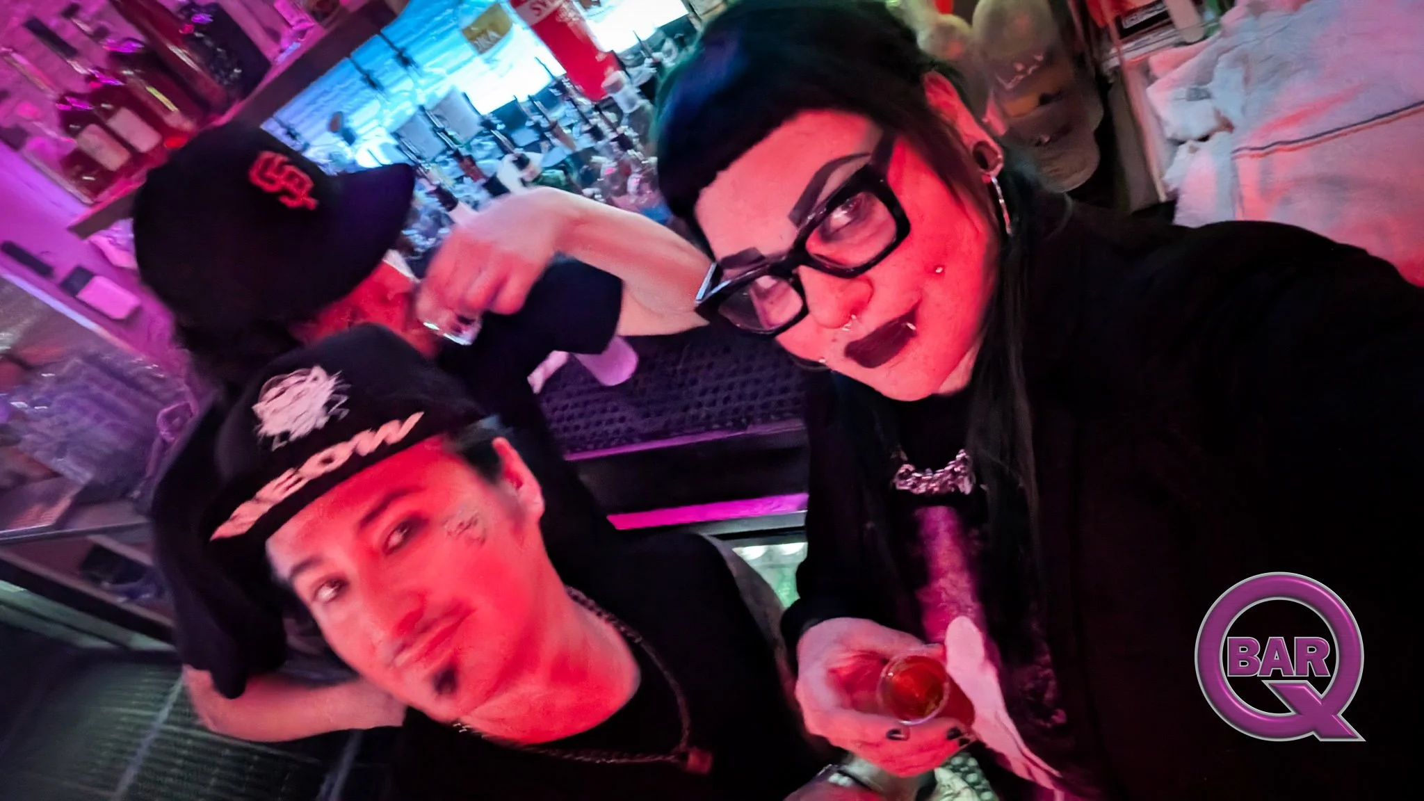 Two people taking a selfie inside a bar. The woman has black hair, glasses, and piercings, and is holding a drink. The man has face tattoos and a bandana. Neon lights and bar shelves with bottles are visible in the background.