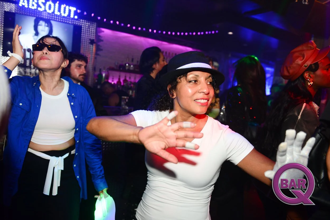 People dancing and enjoying themselves at a nightclub with vibrant purple and pink lighting. A woman in the foreground wears a white t-shirt and a black hat, smiling and dancing with her hands raised. In the background, another woman in sunglasses an