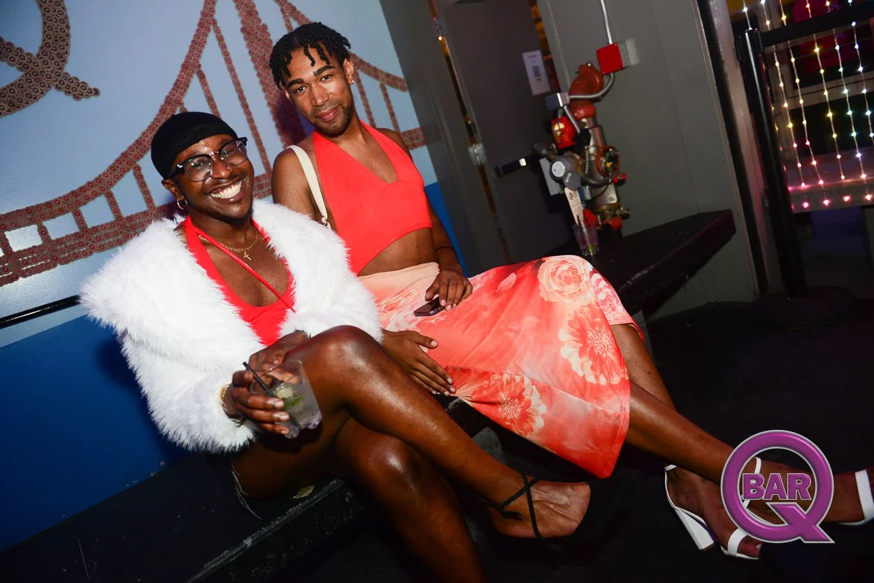 Two men sitting on a bench at a bar, smiling. The man on the left is wearing glasses, a black hat, a white furry jacket, red shirt, and shorts. The man on the right is wearing a coral-colored skirt and coral top. There is a wall art of the Golden Gat