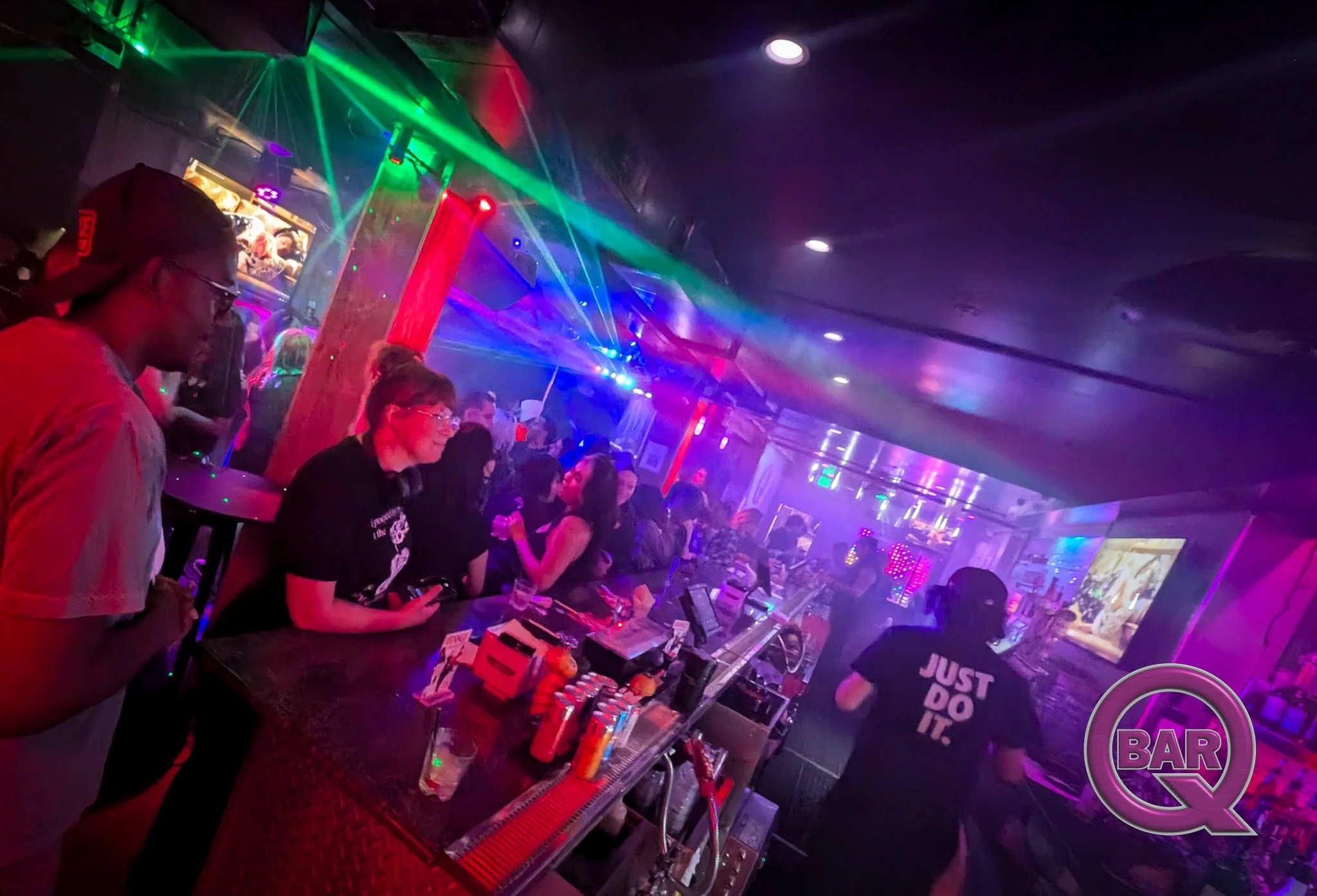 People in a nightclub at a bar with colorful neon lights and laser effects. A woman in the foreground is looking at her phone while others chat and enjoy drinks.