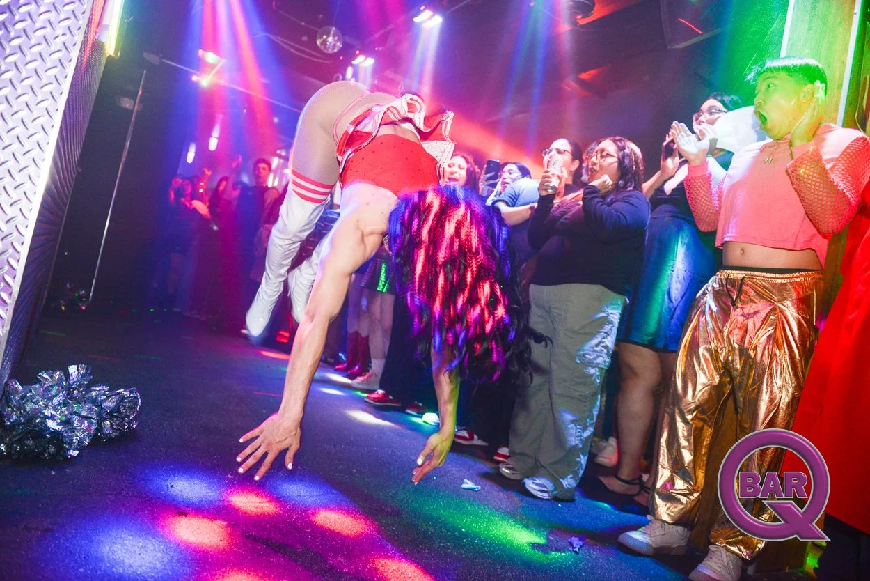 A performer with colorful hair doing a backbend in front of a surprised crowd at a nightclub. Bright, colorful lighting and a 'Bar Q' sign in the corner.