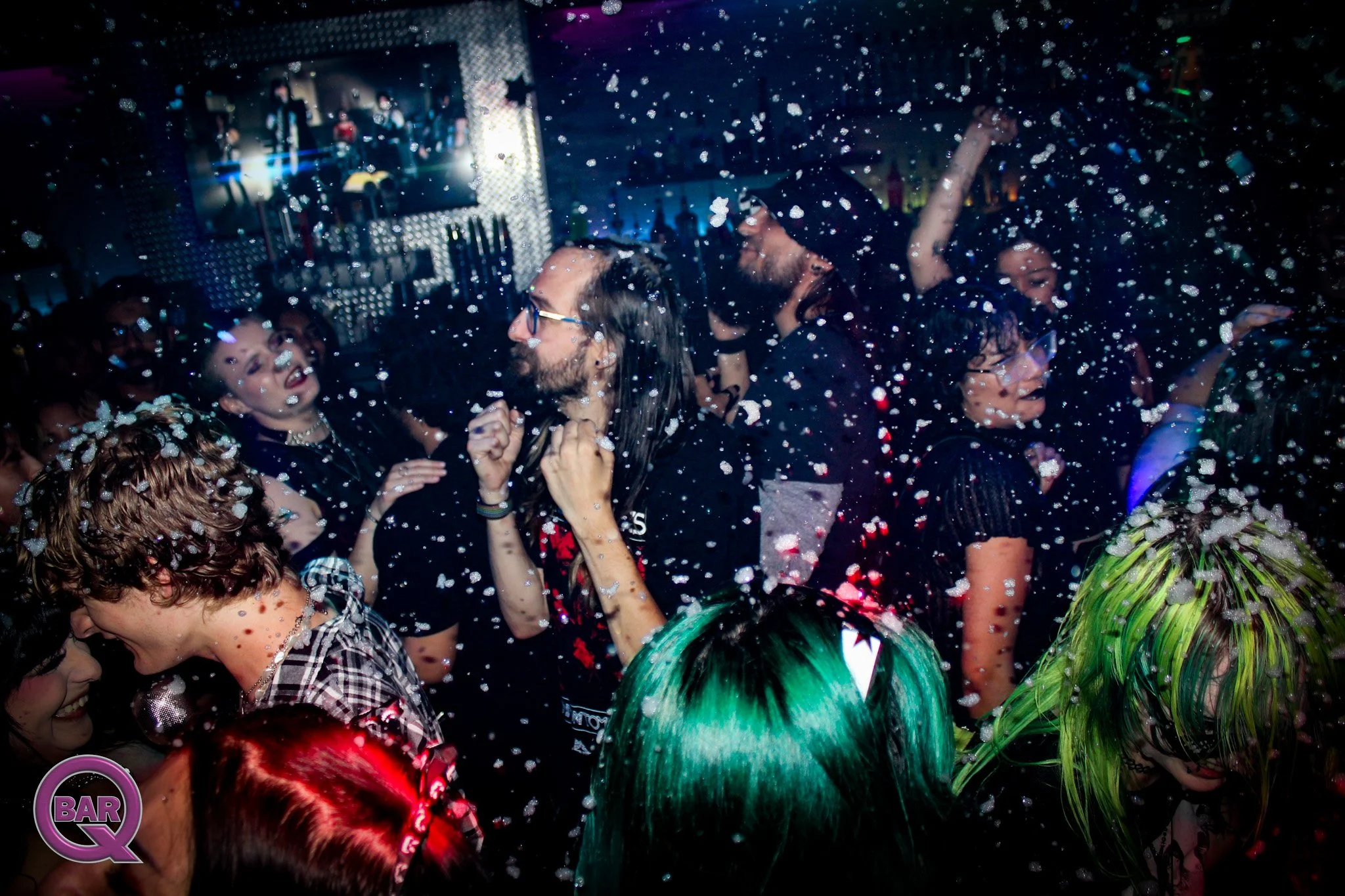 People dancing and celebrating at a party with foam and sparks flying, a large TV screen in the background, and colorful lighting.