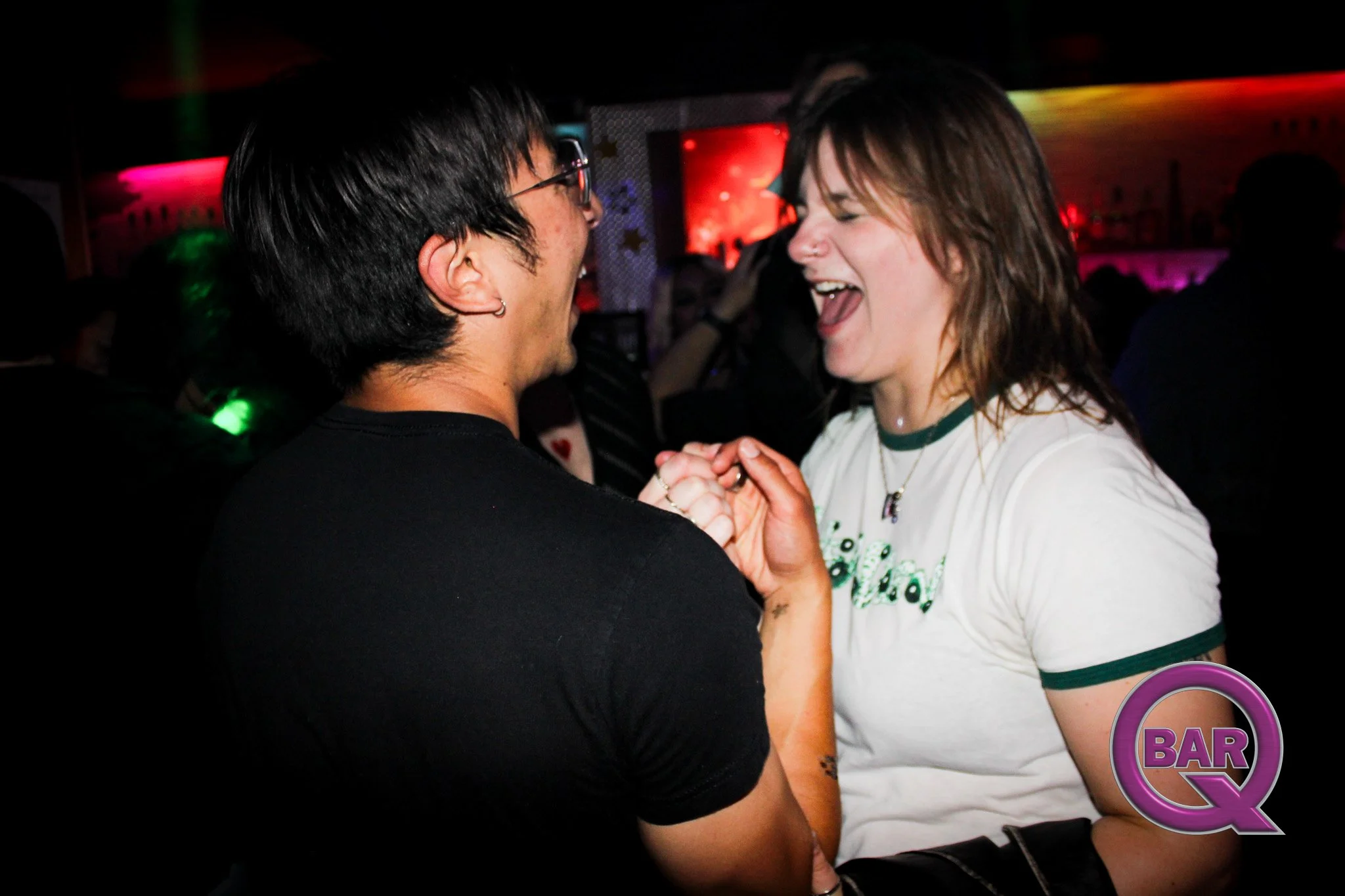 Two people, a man with glasses and a woman, smiling and holding hands in a dimly lit nightclub or bar setting with colorful background lighting.