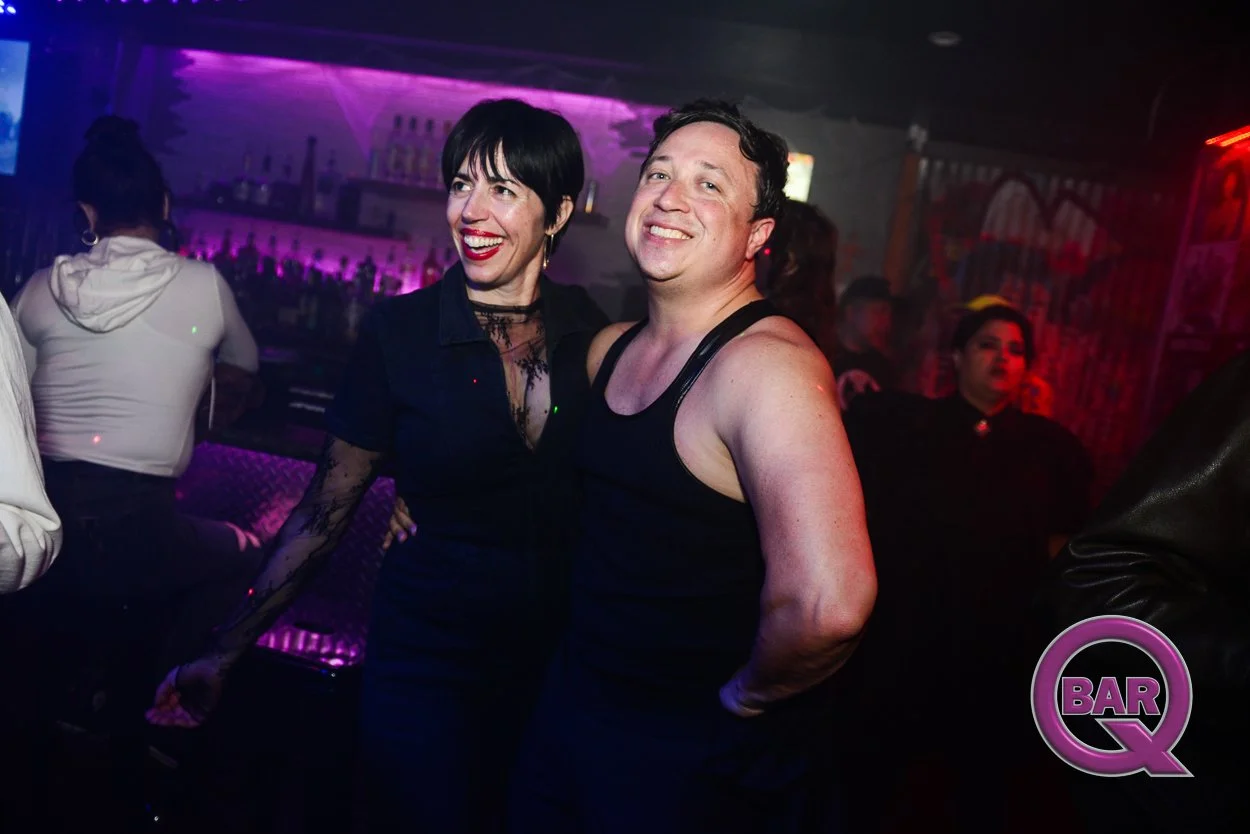 A man and woman smiling and posing together at a bar or nightclub with purple lighting and other people in the background.