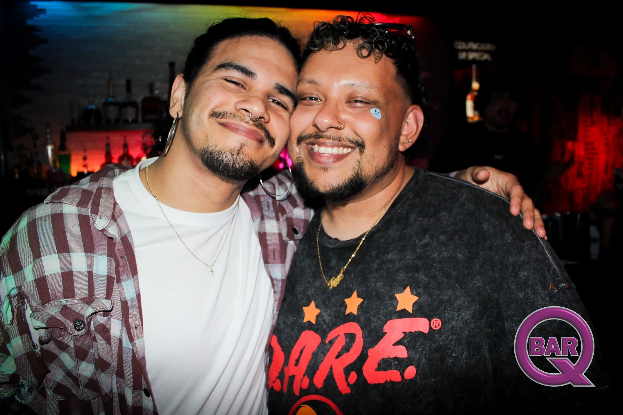 Two men smiling and embracing each other at a bar or club with colorful lighting in the background.