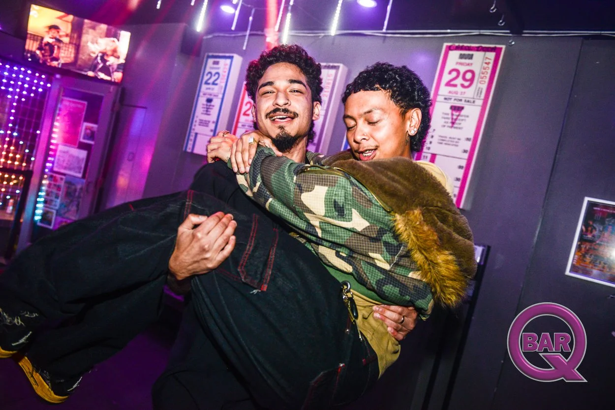 Two young men dancing together at a bar or nightclub with colorful neon lights and posters on the wall behind them.