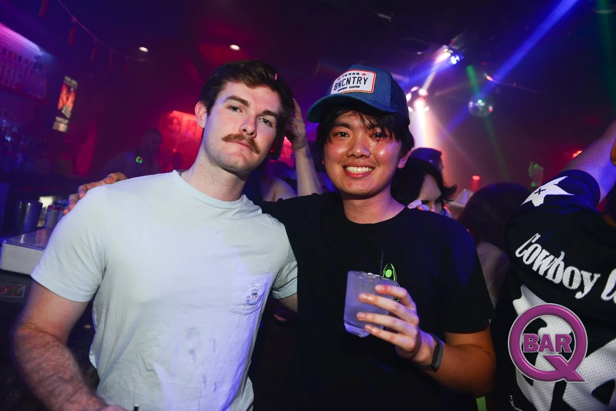 Two men posing together at a nightclub with colorful lights, one wearing a white t-shirt and the other wearing a black shirt with a baseball cap, holding a drink.