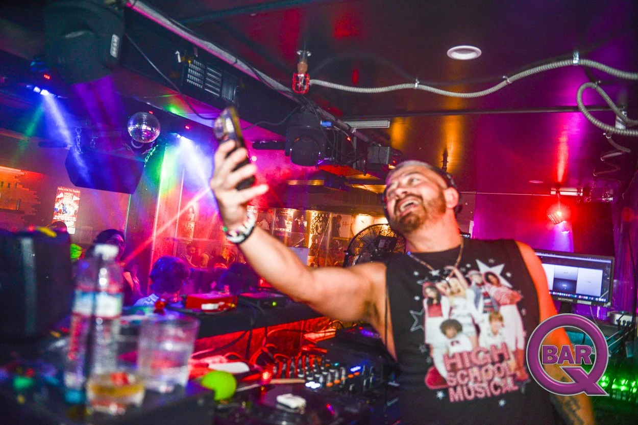 A male DJ with a beard and mustache smiling and taking a selfie in a colorful, vibrant nightclub with purple, green, and red lighting. The DJ wears a black sleeveless T-shirt with a cartoon graphic and the words 'High School Music.' The DJ's equipmen