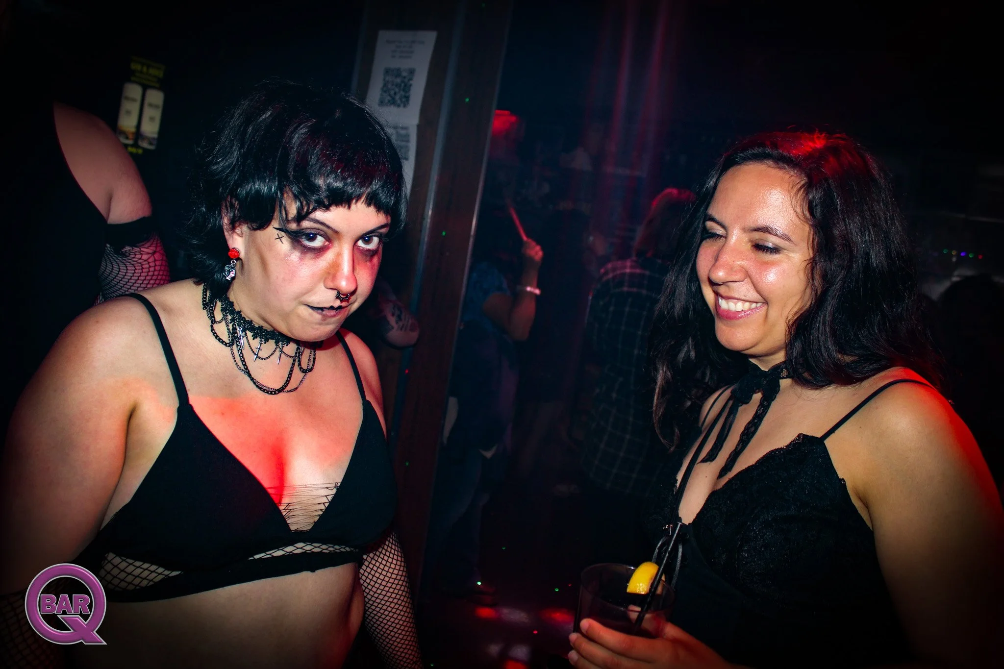 Two women dressed in black lingerie at a nightclub, with one showing makeup and jewelry, and the other smiling while holding a drink with a lemon slice and straw.