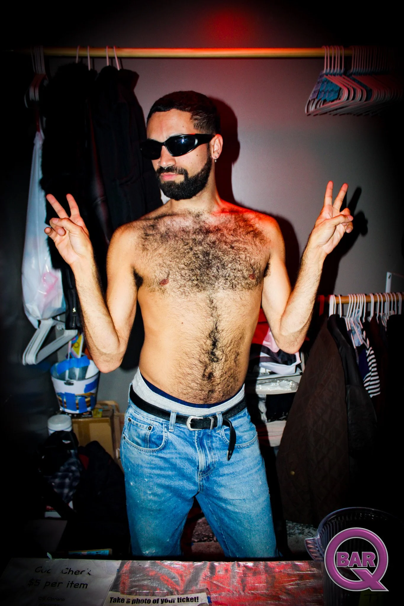 A shirtless man with long hair, beard, and sunglasses making peace signs with both hands in a closet or dressing room with hangers and clothing in the background.