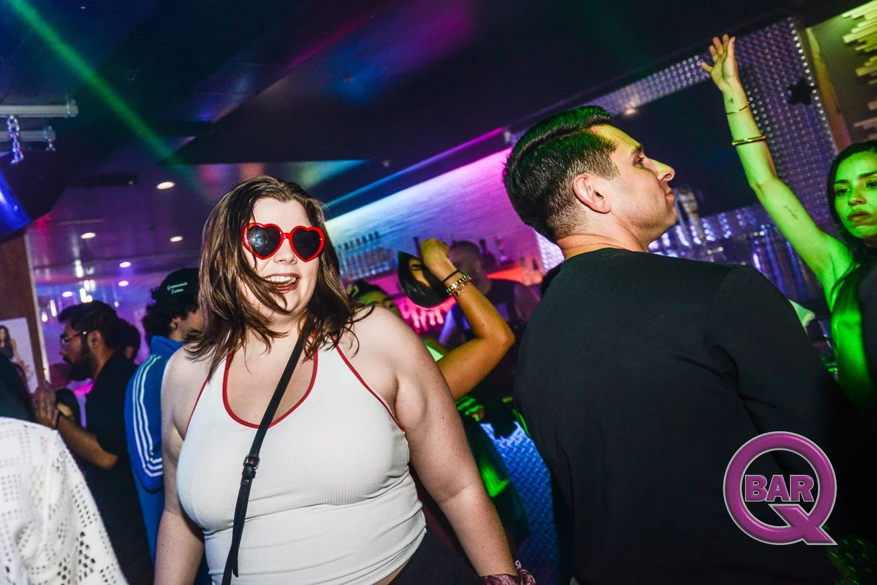 A woman wearing heart-shaped red sunglasses and a white tank top with red trim smiling in a crowded, colorful nightclub with neon lights, other dancing people, and a bar in the background.