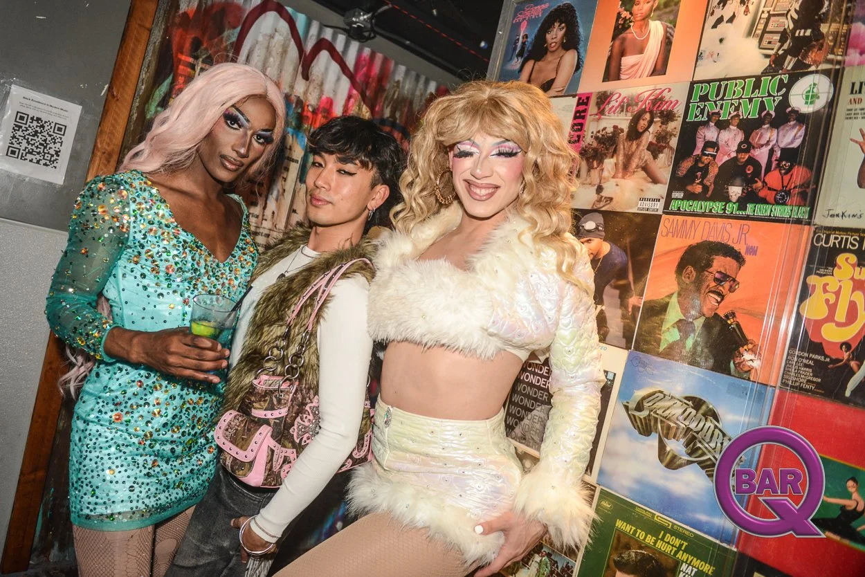 Three drag queens standing together in a room decorated with music album covers and posters on the wall.