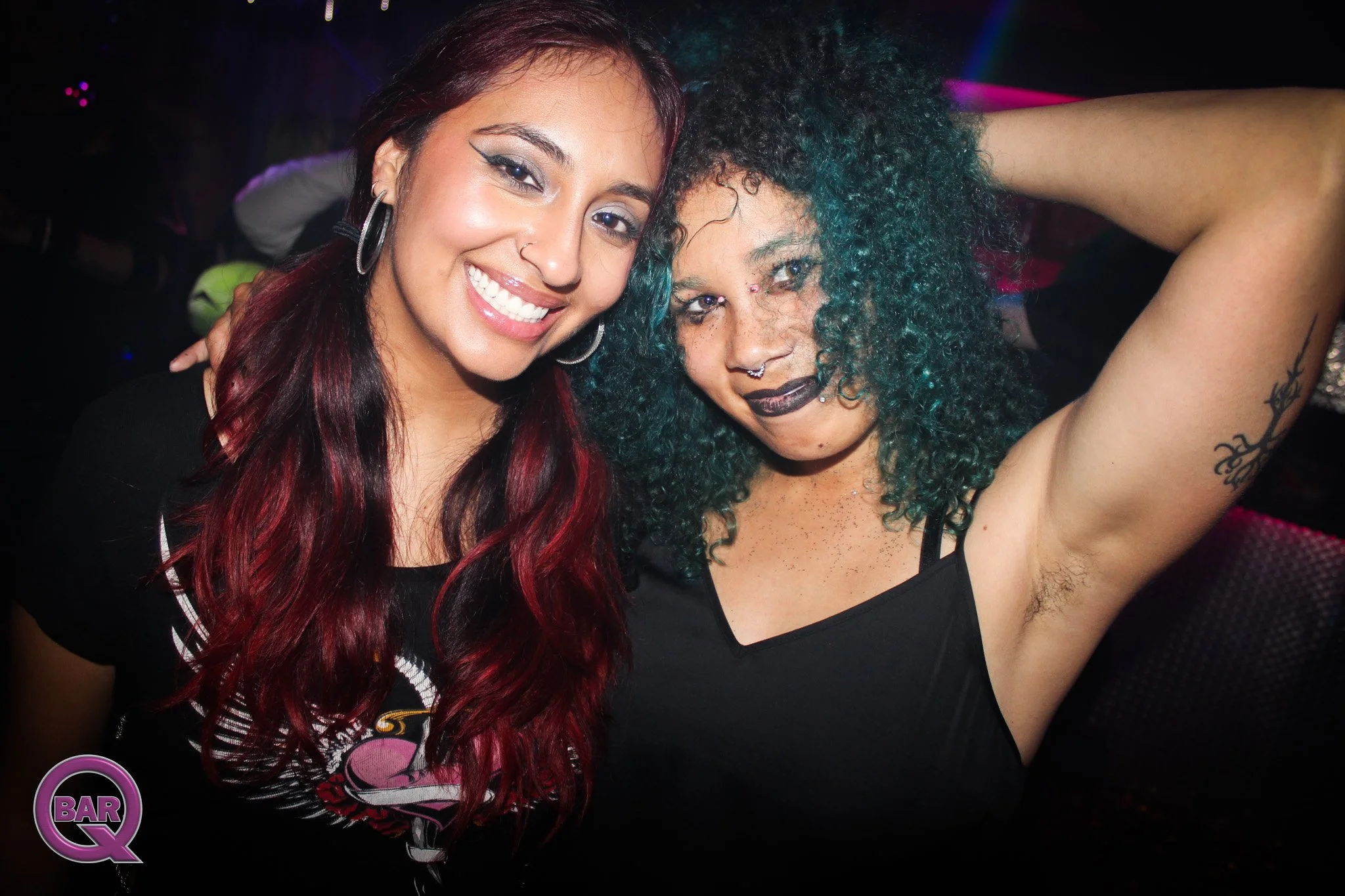 Two women smiling for a selfie in a nightclub. One has red hair and the other has curly teal hair. Both are wearing black clothing and have facial piercings.