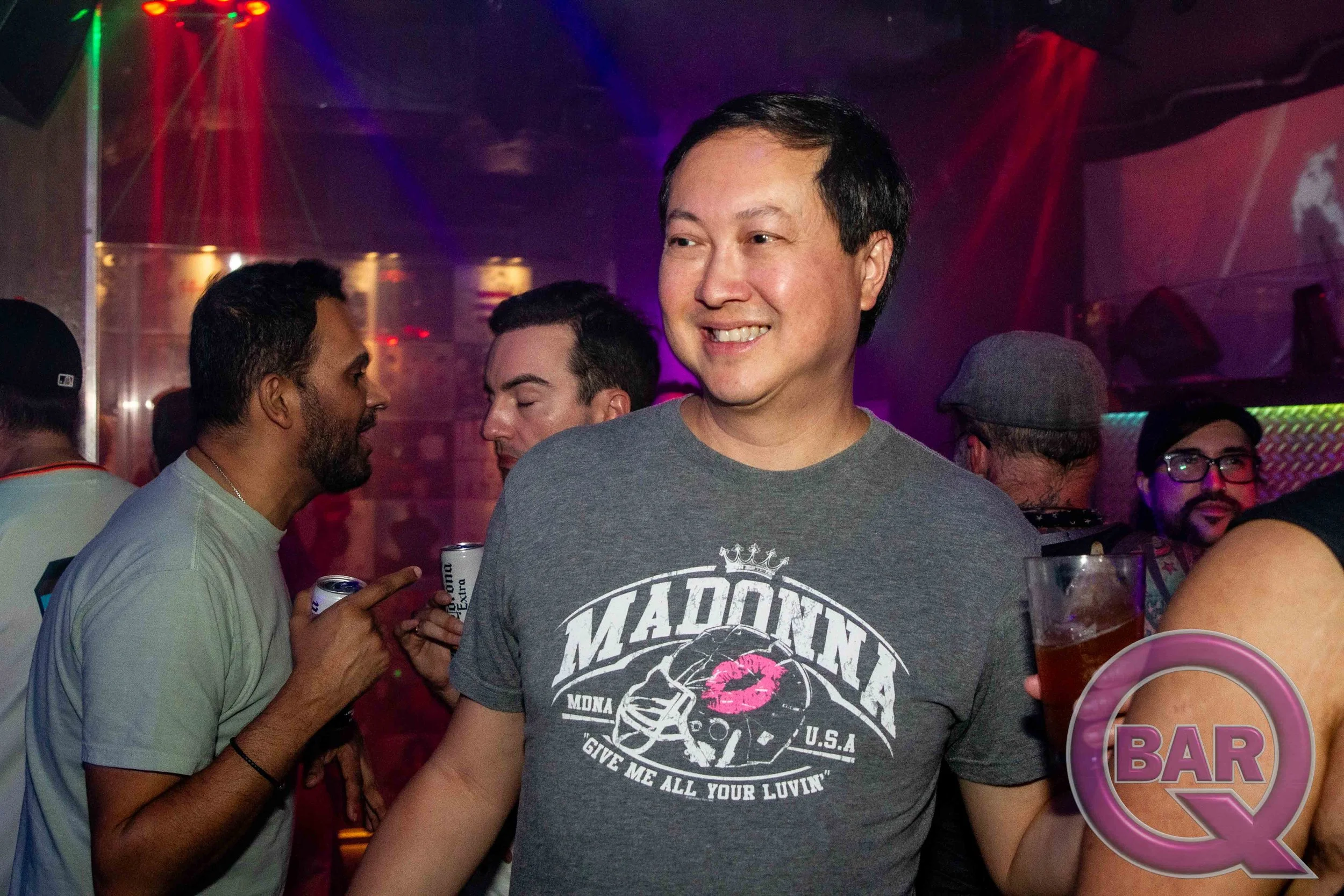 A group of people socializing at a bar or nightclub with colorful lighting, including a man smiling in the foreground wearing a gray T-shirt with a graphic design.
