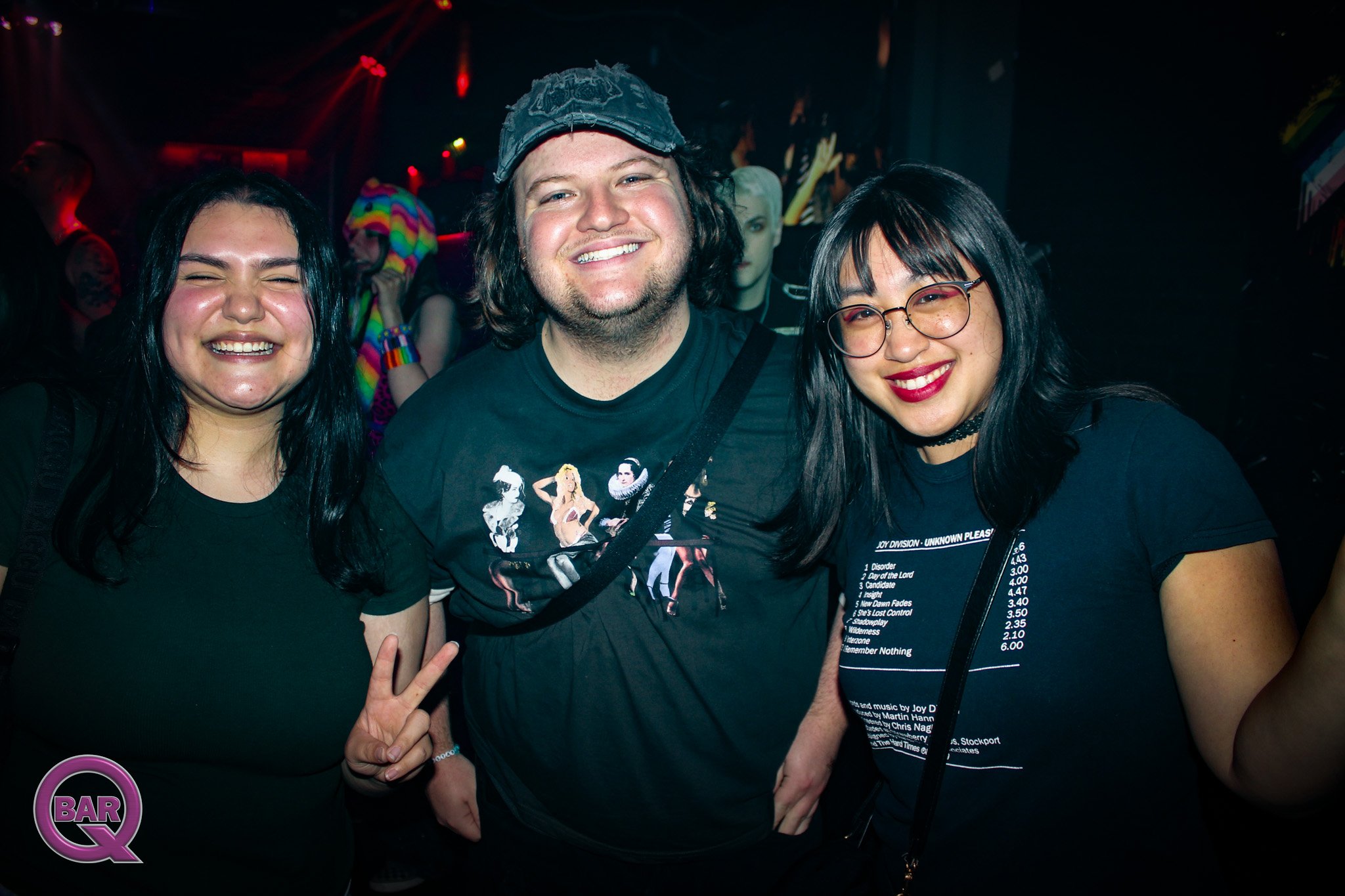 Three smiling people posing in a nightclub, two women and one man, with colorful lights in the background. The woman on the left has long black hair, the man in the middle is wearing a cap, and the woman on the right has glasses and dark hair.