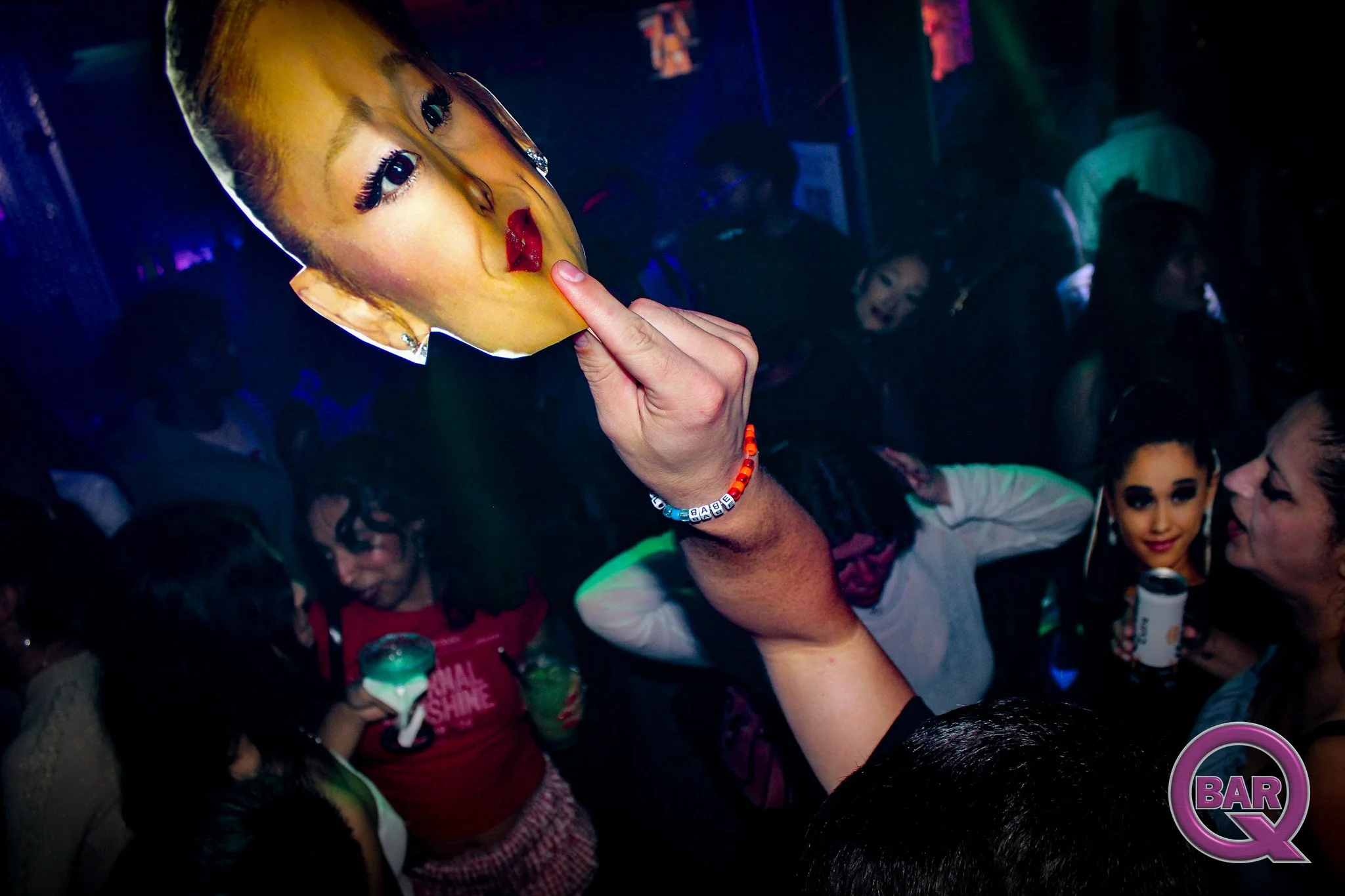 A person holds a paper mask of a woman's face with red lipstick to their face, surrounded by a crowd at a party or club with dim lighting and neon accents.