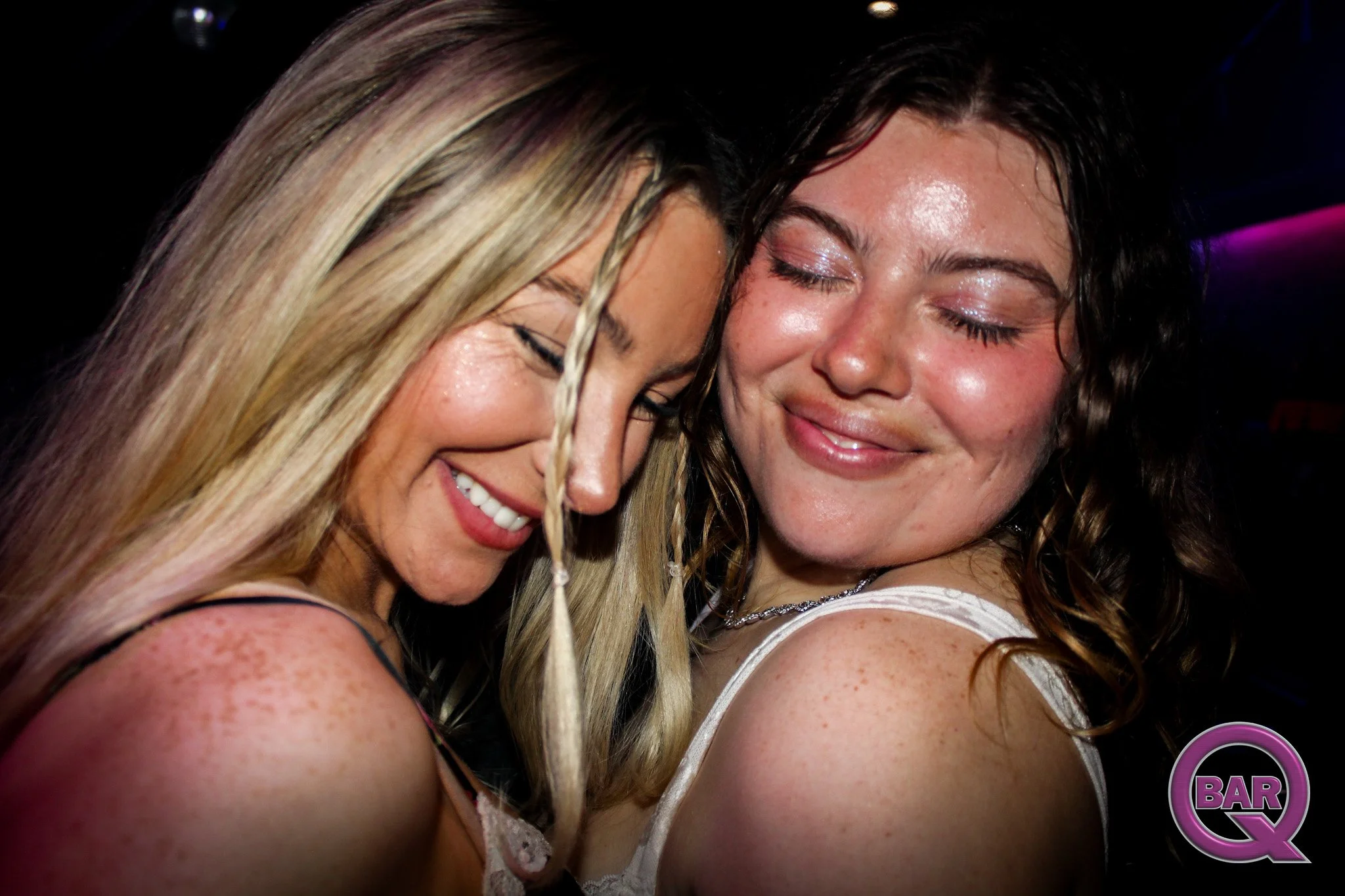 Two women are close together, smiling with their eyes closed, at a cheerful social event or nightclub. One has blonde hair with a braid and the other has dark brown, curly hair. There is a pinkish-purple light in the background.