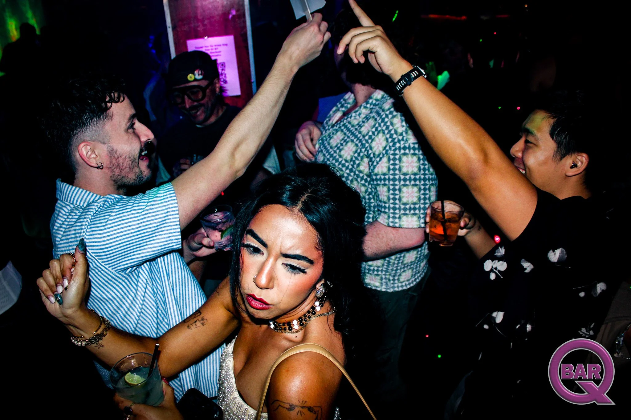 A group of people dancing and socializing at a nightclub, with colorful lights and a bar sign visible in the background.