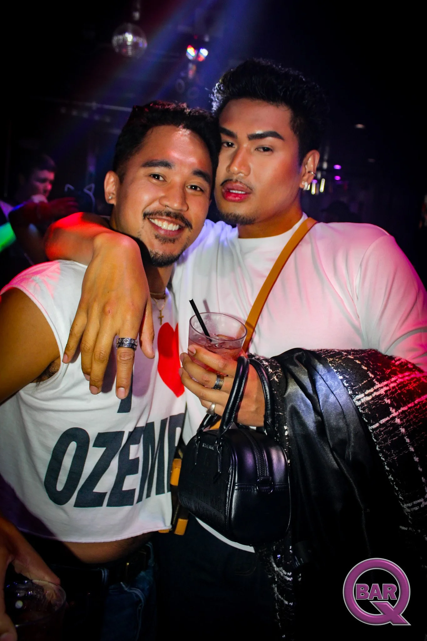 Two men at a bar with the DJ in the background, one holding a drink and wearing a white T-shirt, the other with a black bag, both smiling and posing for a photo.