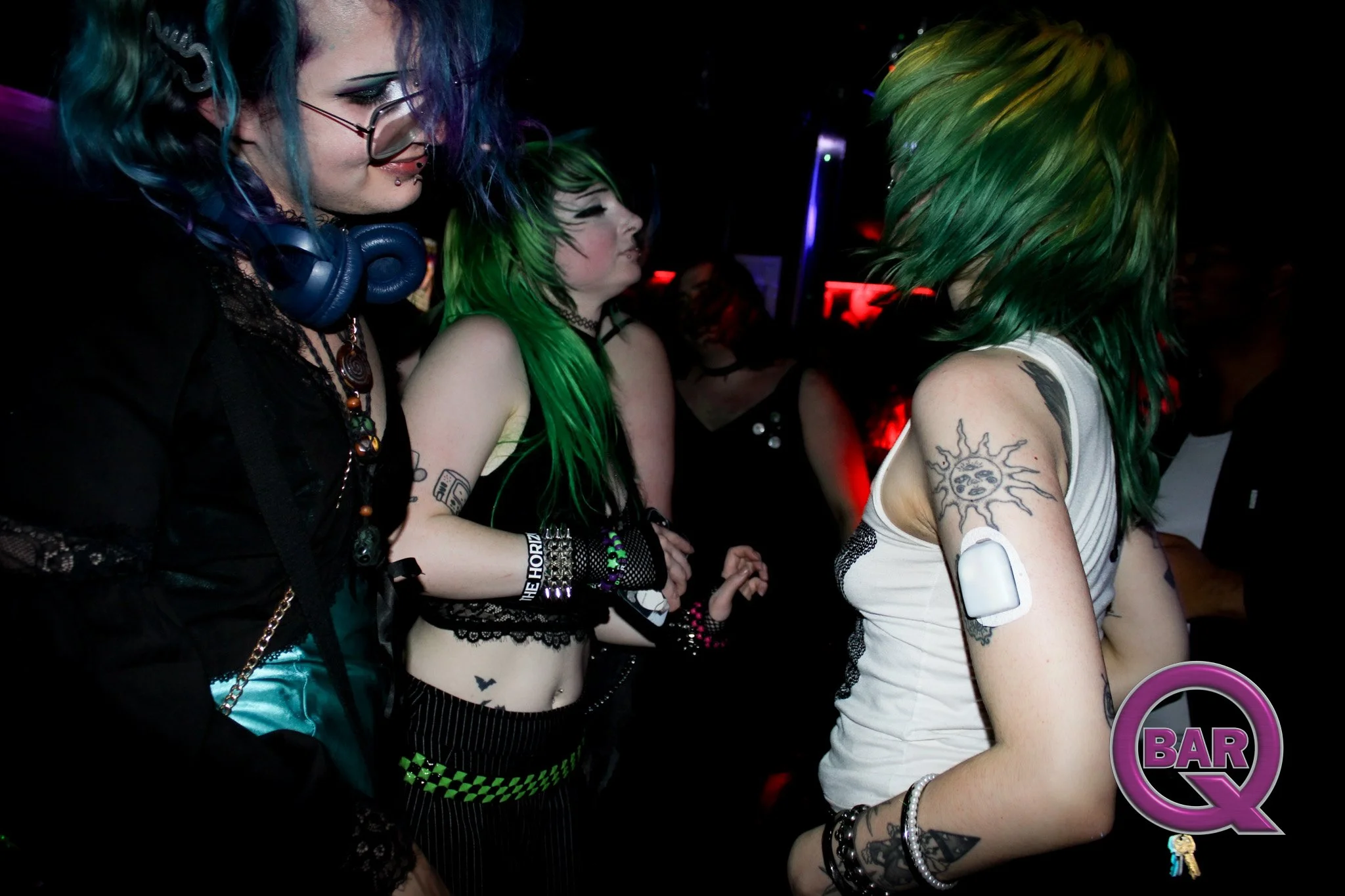 Three women with colorful hair and tattoos standing in a dark bar or club, engaged in conversation.