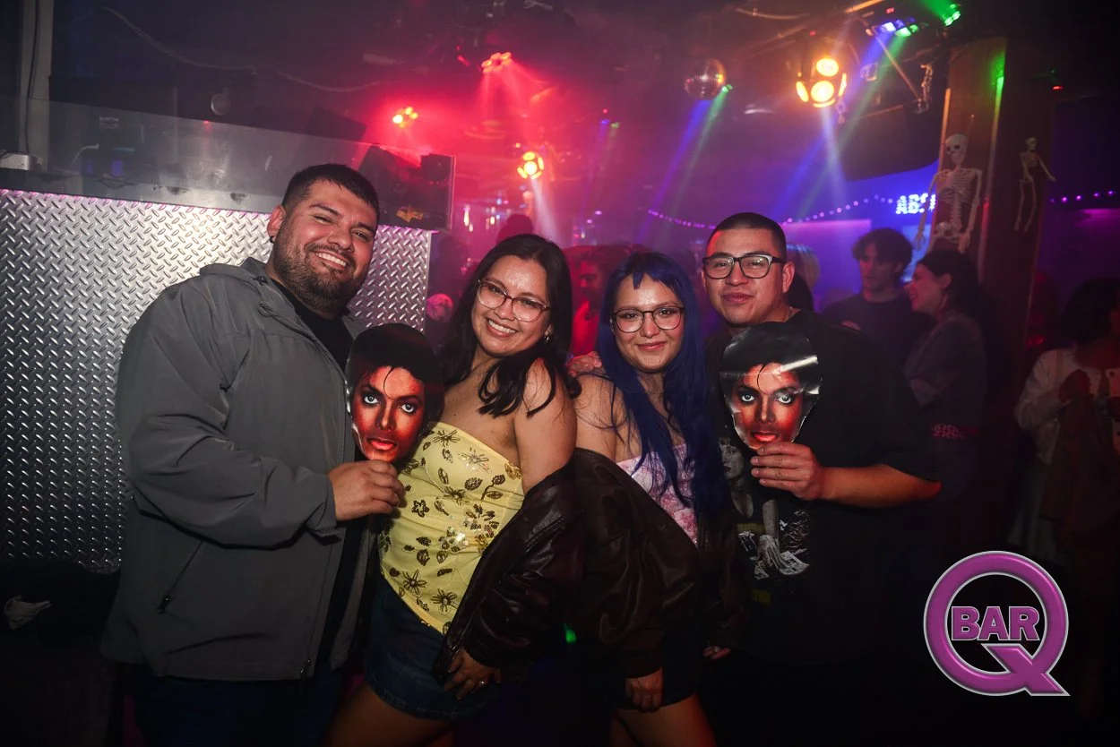Group of four friends smiling and holding images of Michael Jackson at a nightclub with colorful lights and skeleton decorations in the background.