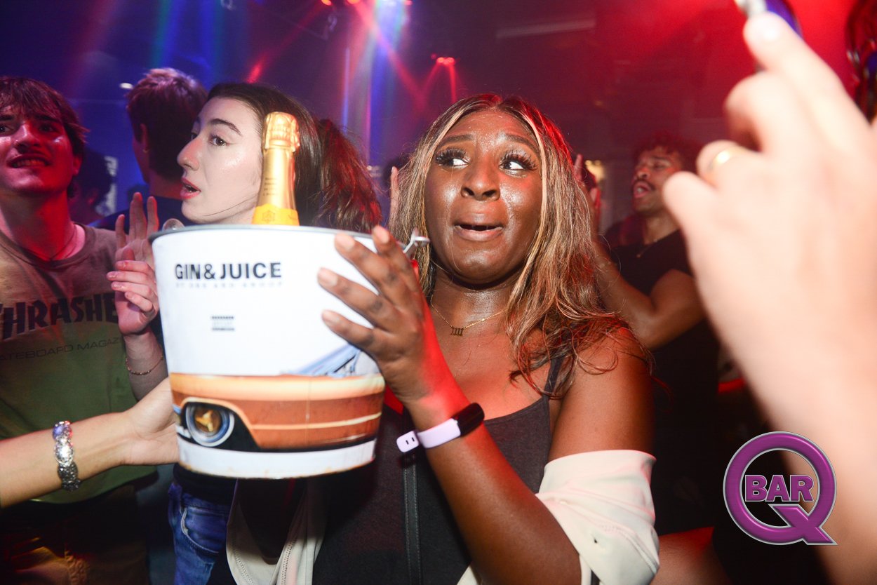 A woman at a bar surrounded by friends, holding a bucket with a bottle of champagne, a bottle of gin and juice logo visible, colorful dance party lighting, some people looking around and enjoying the night.