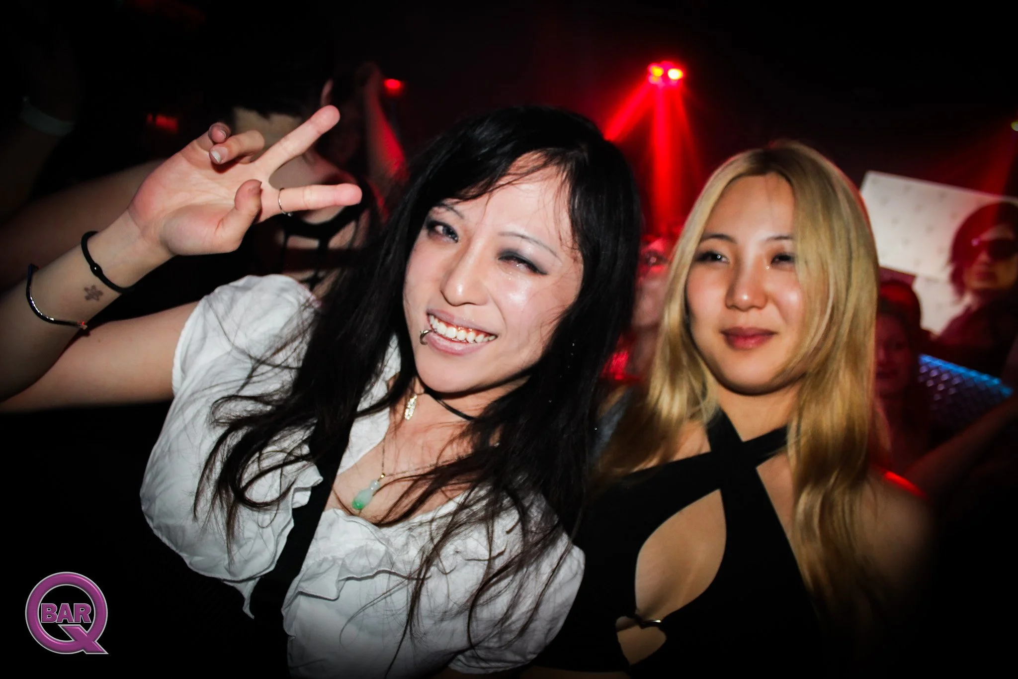 Two women dancing at a nightclub with red lights in the background. One woman has black hair and is making a peace sign, the other has blonde hair. A purple and black logo with the text 'BAR Q' is visible in the bottom left corner.